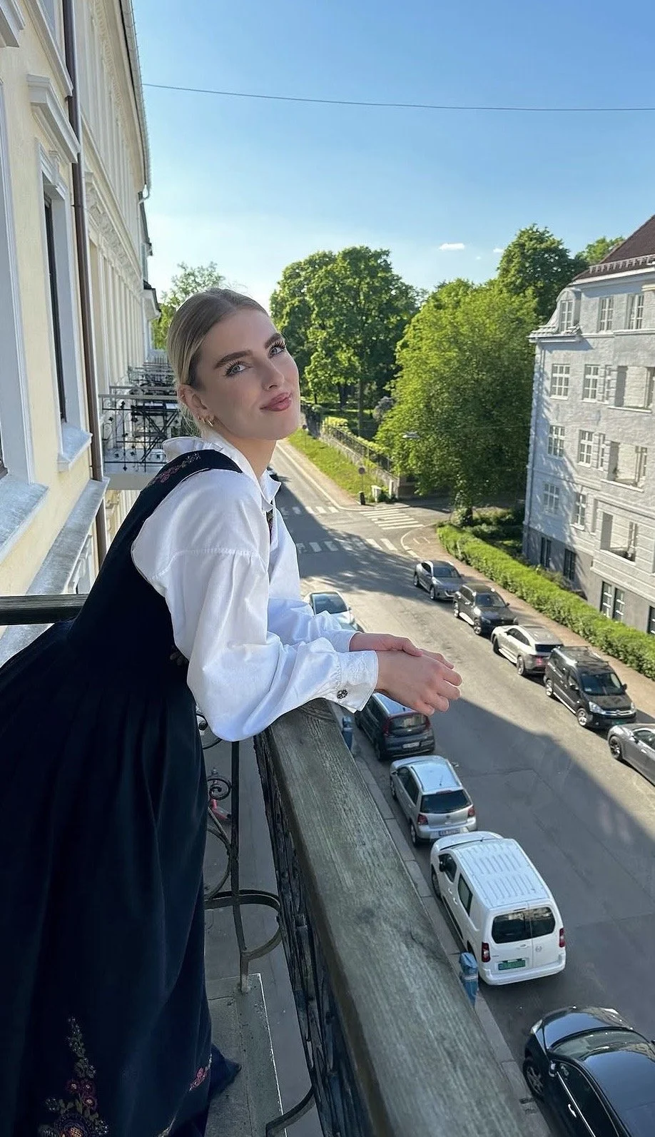 Young woman with blonde hair in a bun, wearing a white shirt and black dress, leaning on a balcony railing, overlooking a street with parked cars and green trees under a clear blue sky.