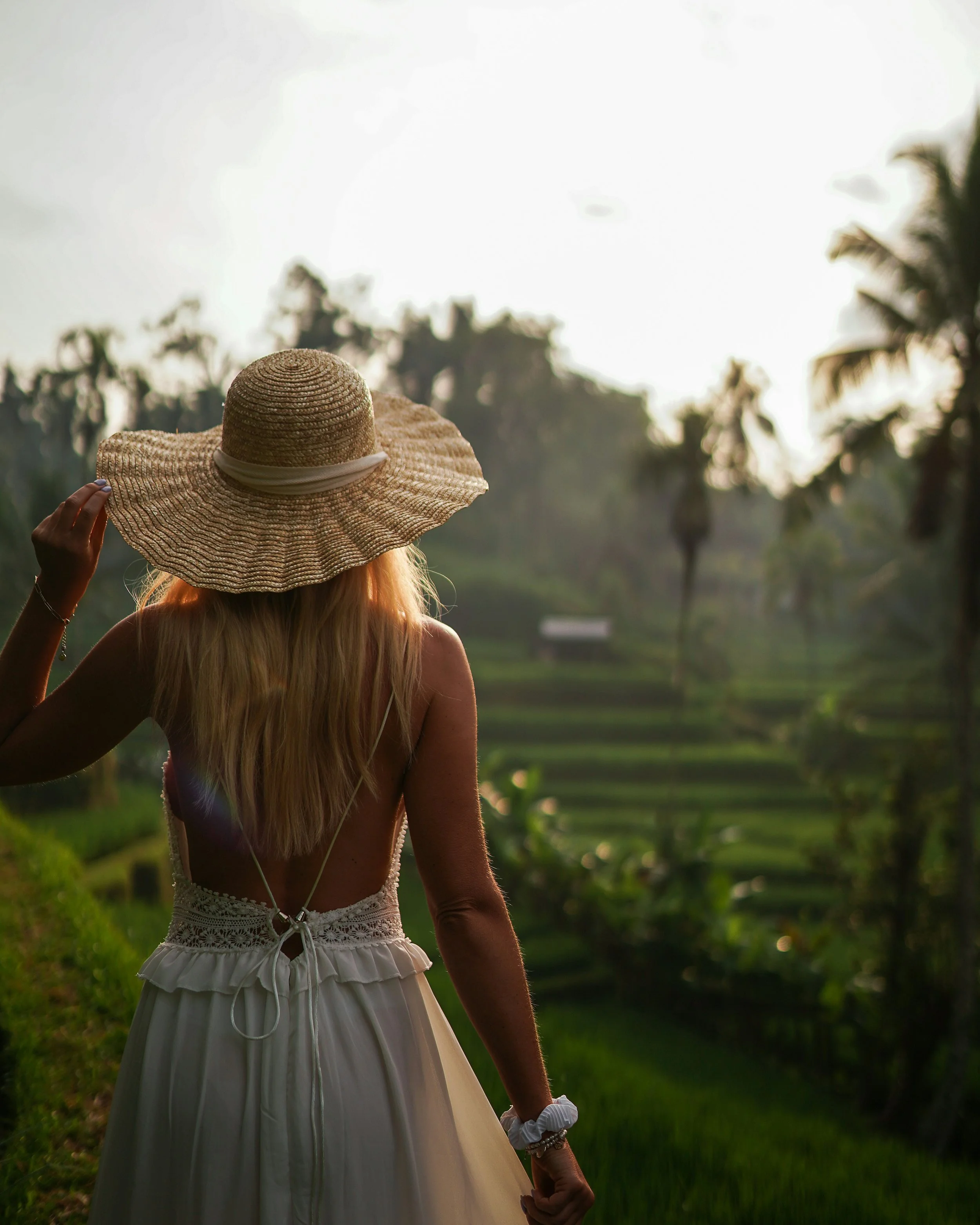 A woman holding a wide-brimmed straw hat with a white band, standing outdoors in a lush green landscape with trees and rye grass, in the late afternoon or early evening.