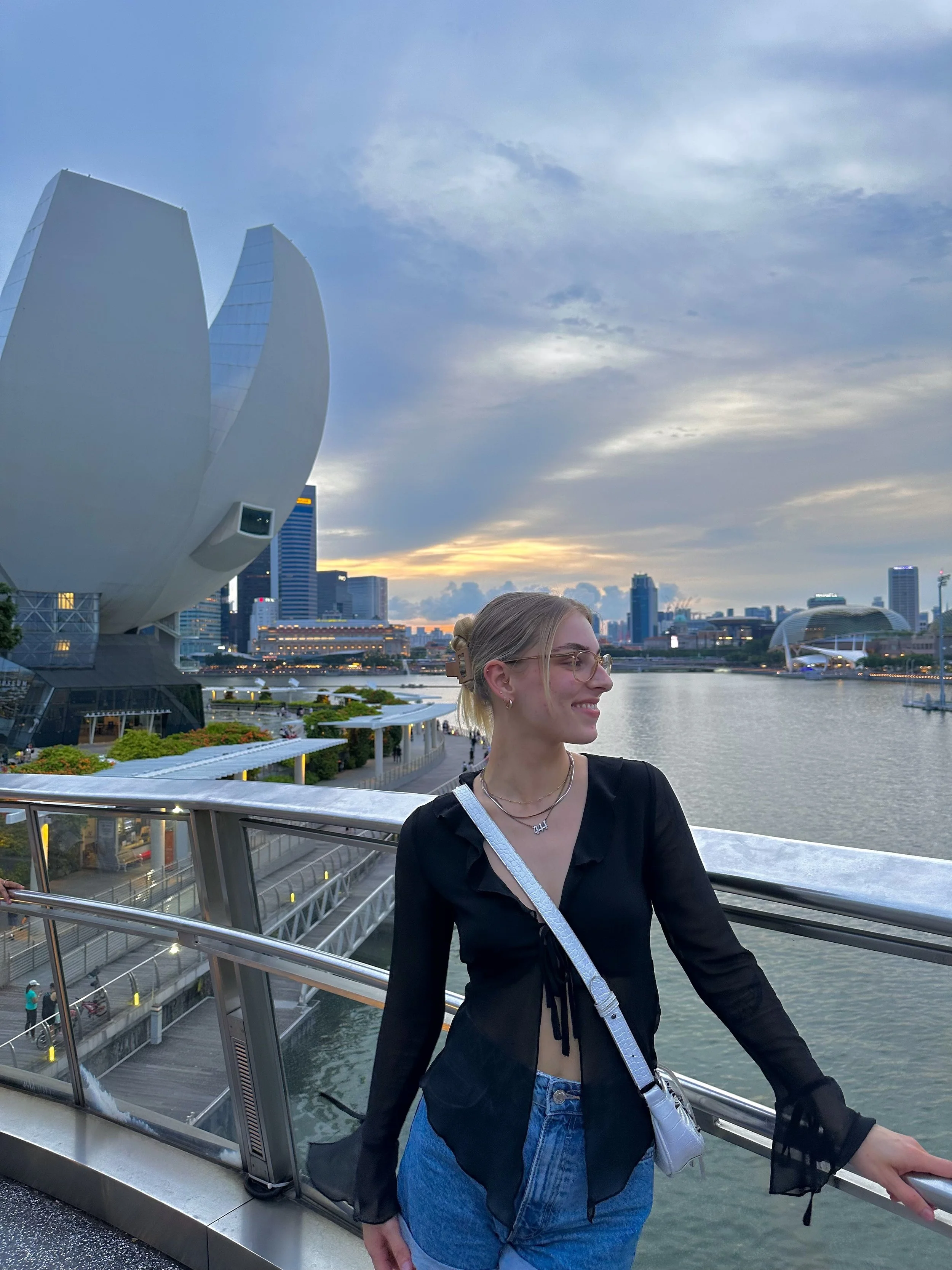 A woman smiling, wearing glasses, a black top with sheer long sleeves, and blue jeans, standing on a bridge overlooking a river with a city skyline and modern buildings in the background during sunset.