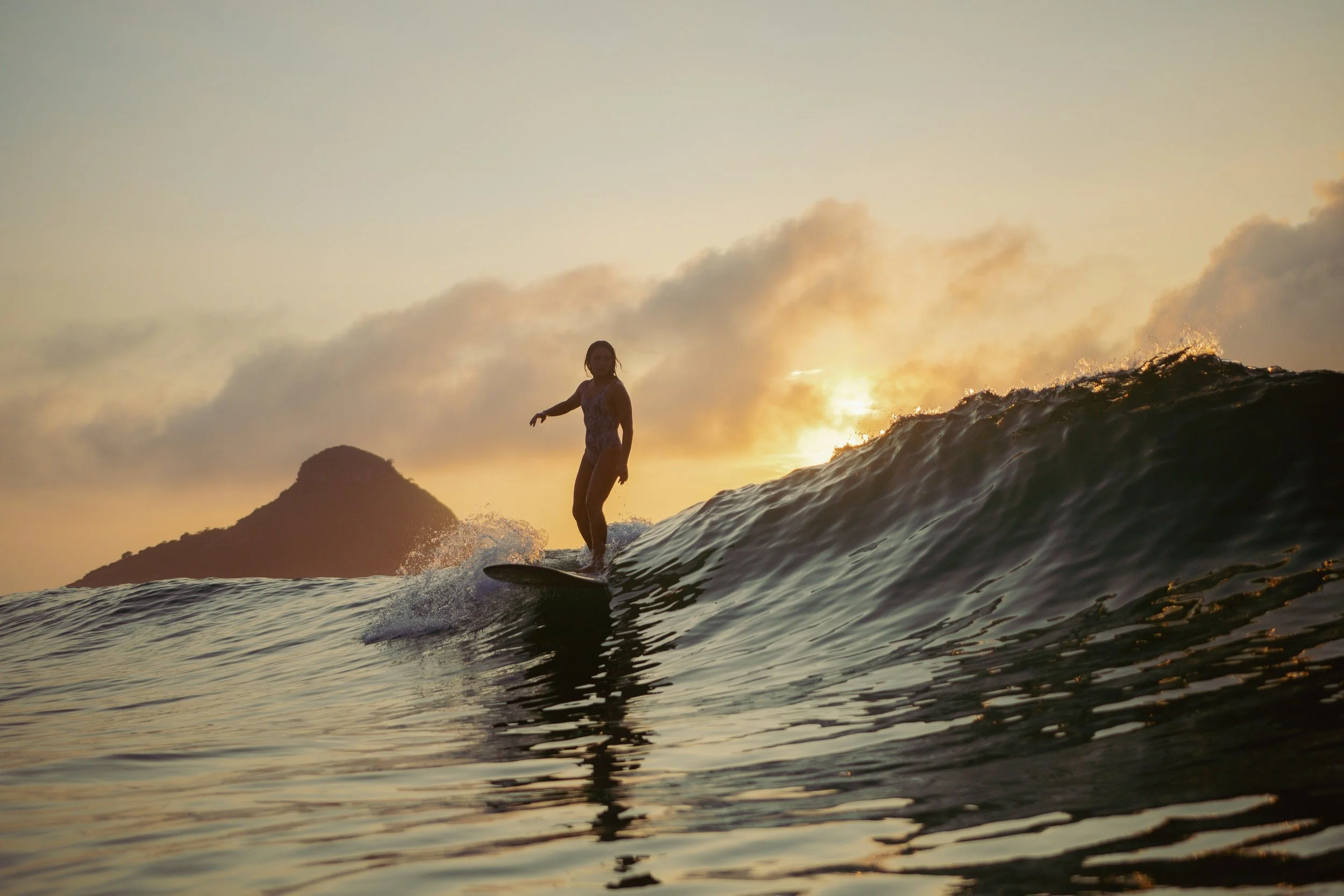 A person surfing on a wave during sunset with mountains and an orange sky in the background.