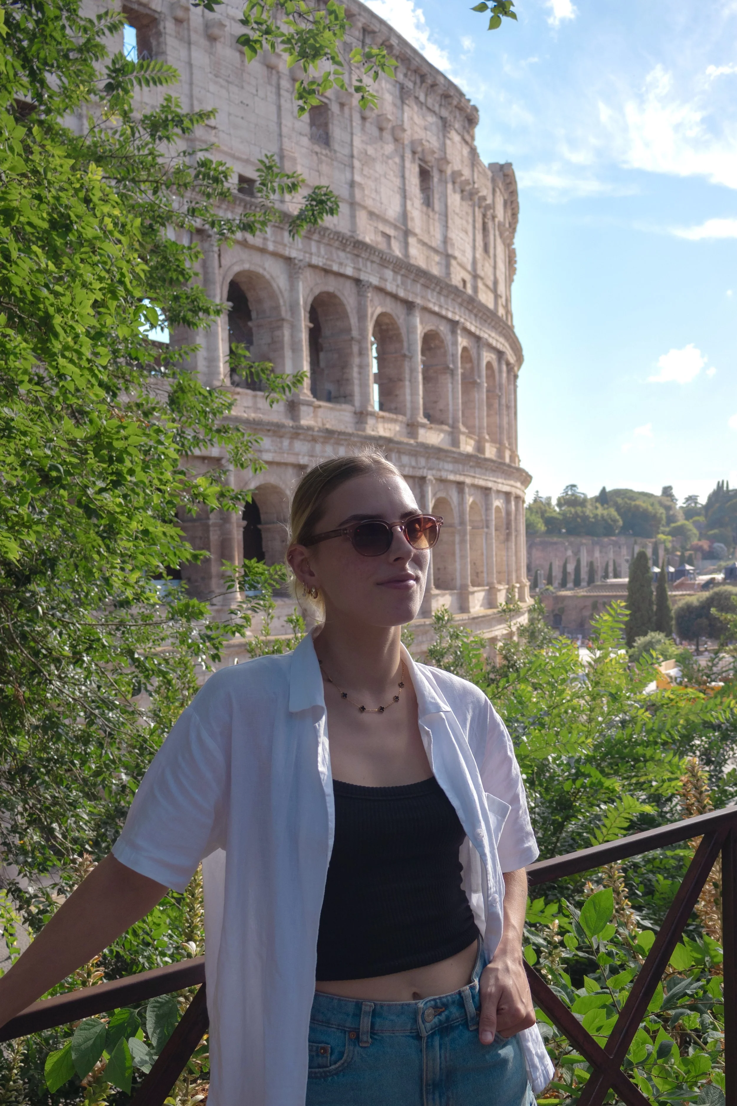 A woman wearing sunglasses, a white open shirt over a black crop top, and jeans, standing outdoors near green foliage, with the Colosseum in Rome visible in the background.