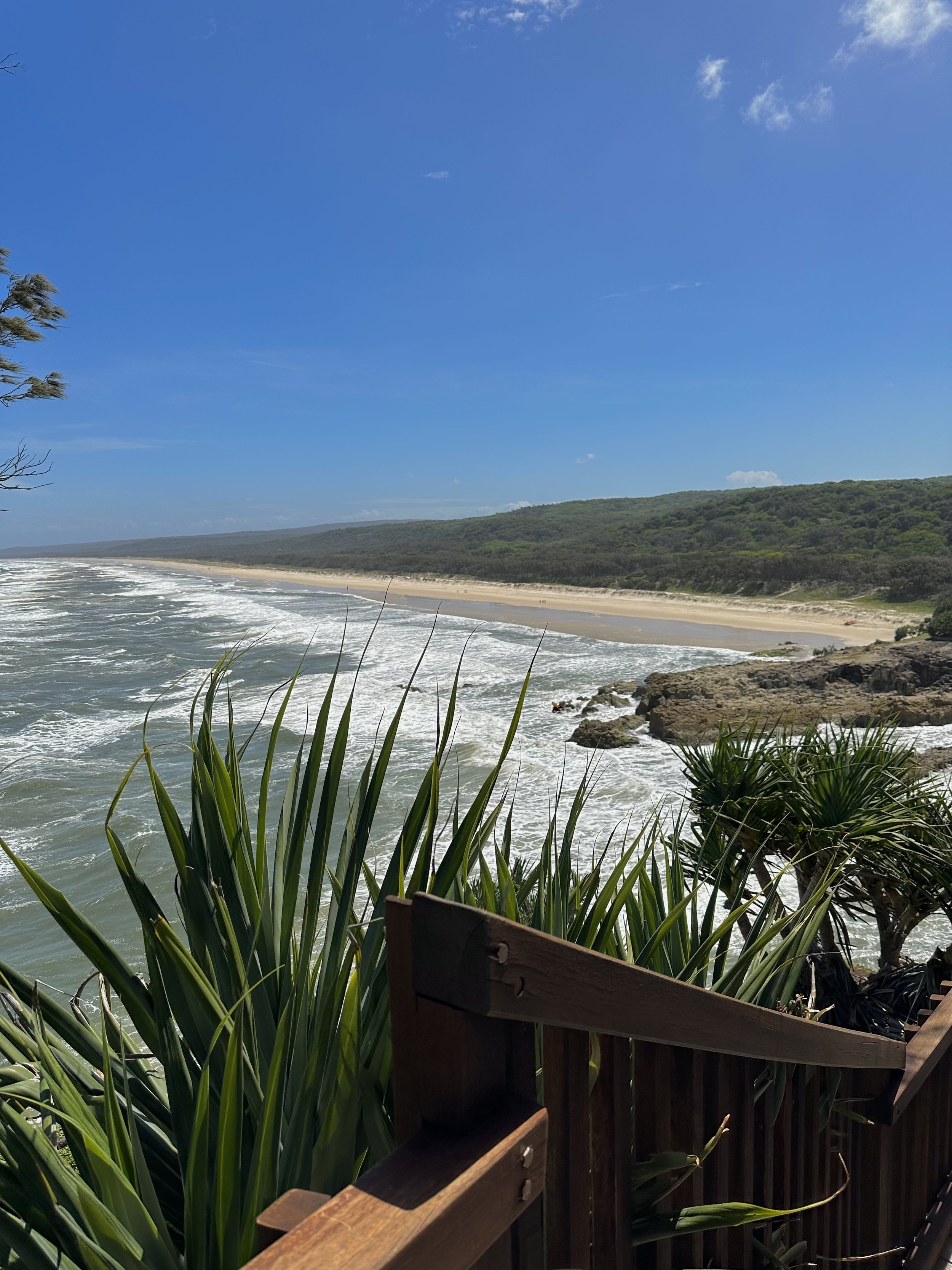 View of a beach with sandy shore, ocean waves, and a green hillside in the distance under a blue sky with a few clouds.