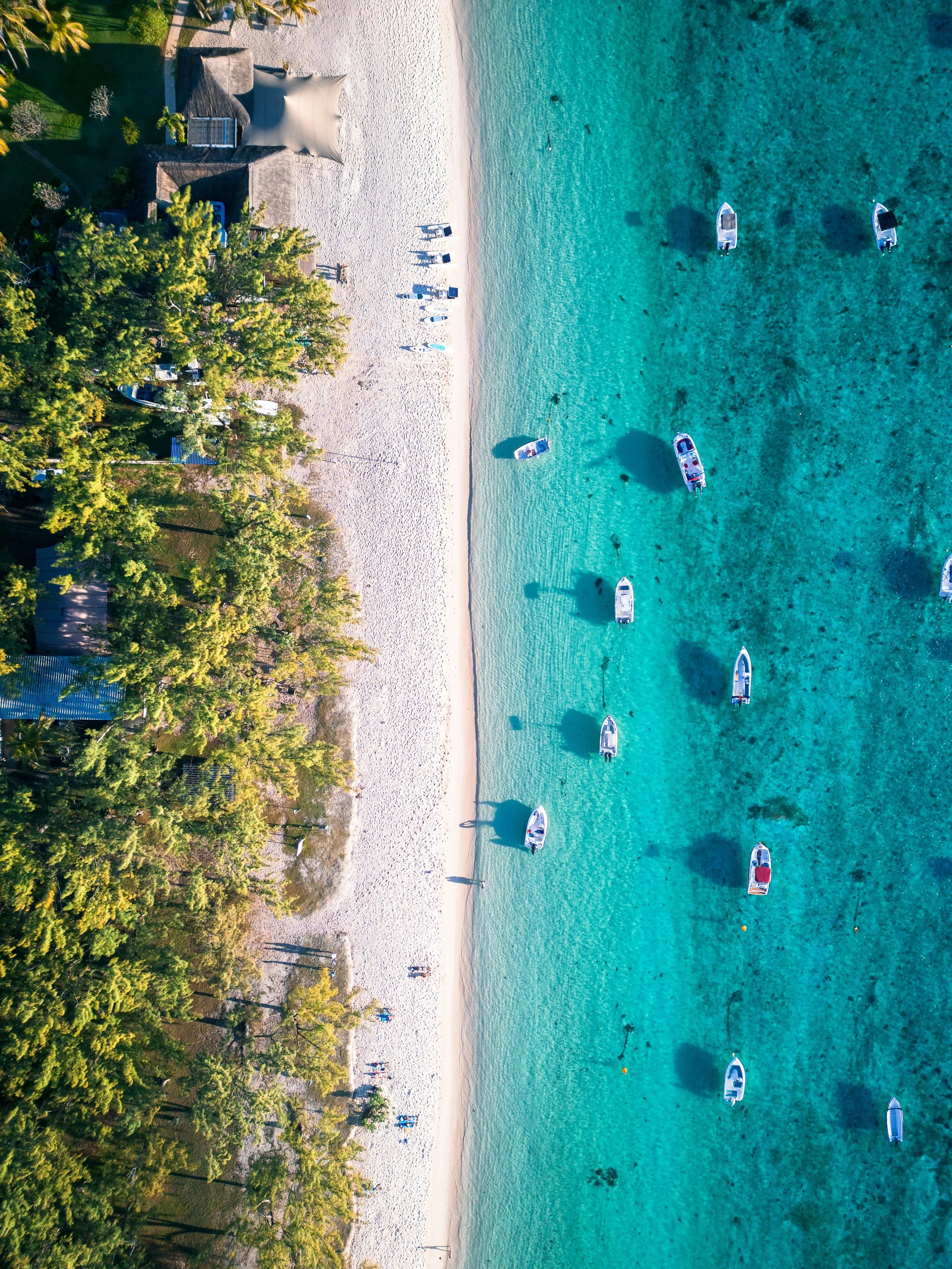 Aerial view of a beach with clear turquoise water, a white sandy shore, boats anchored in the water, lush green trees along the shore, and people walking and relaxing on the beach.