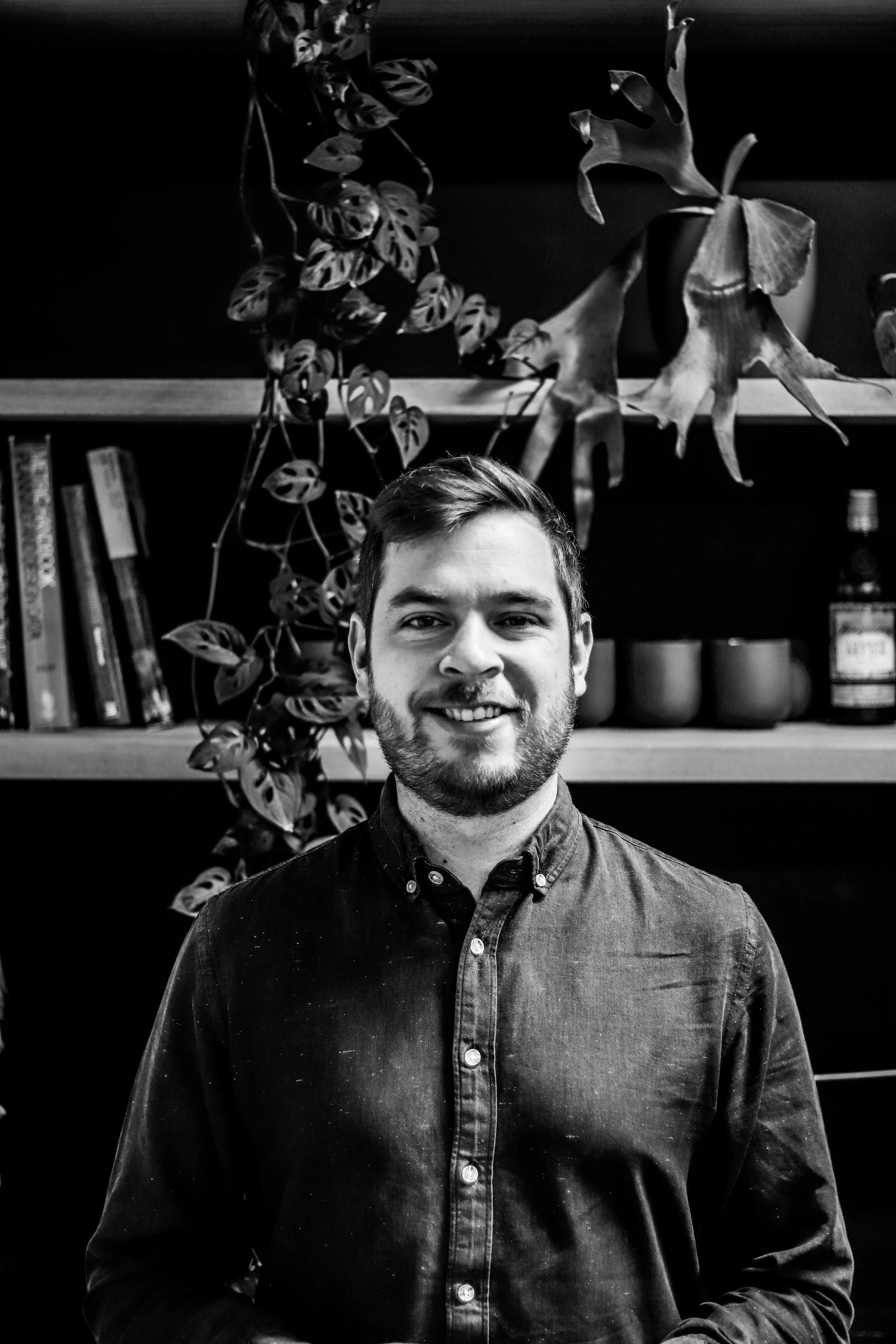 A smiling man with a beard, wearing a button-up shirt, standing in front of a bookshelf with plants and decorative objects in the background.