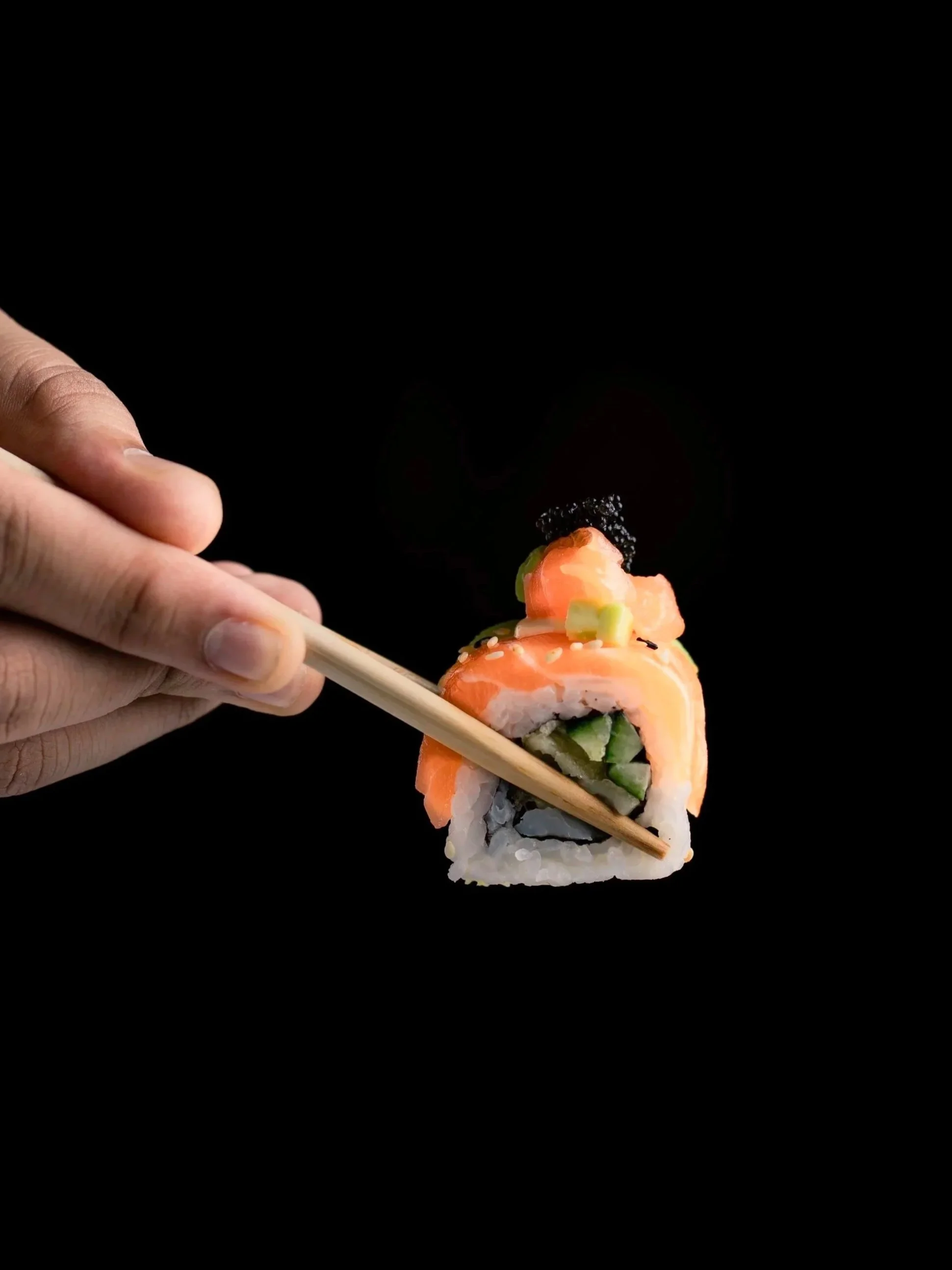 Close-up of a hand holding a piece of sushi with salmon, cucumber, and black caviar on top, against a black background.
