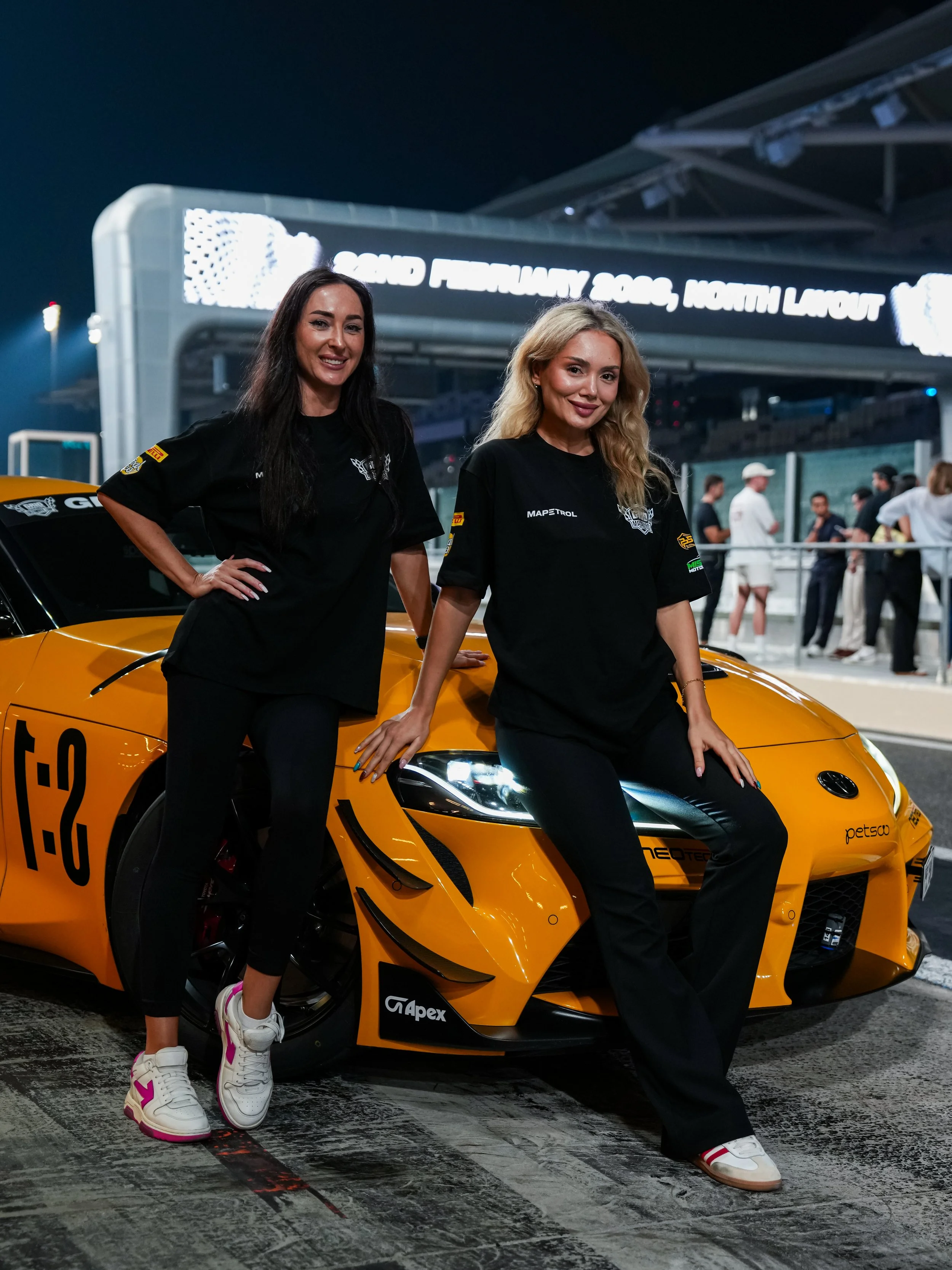 Two women pose next to a yellow sports car at night, with a racetrack and a large digital sign in the background.