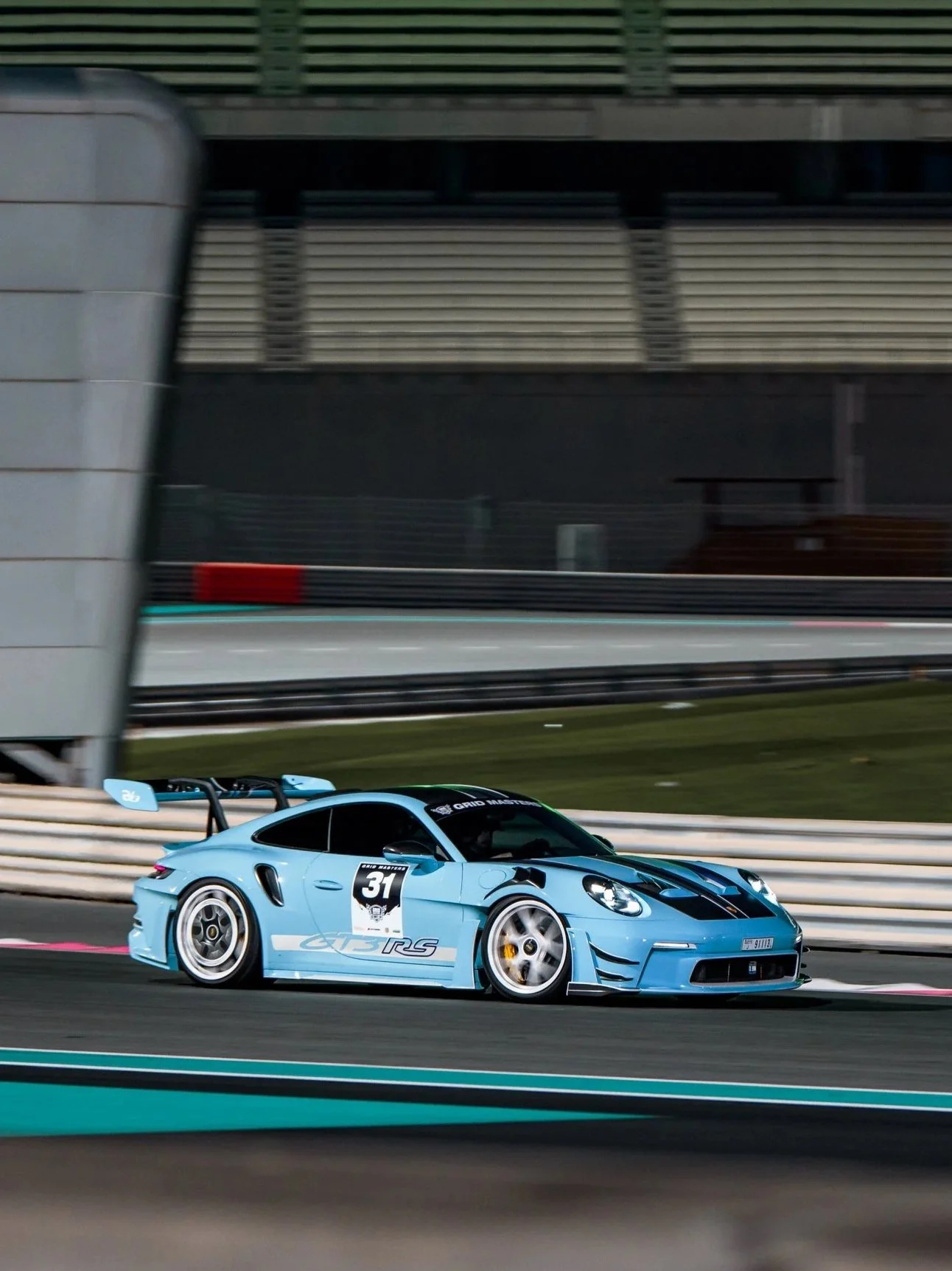 A light blue race car with the number 31 on the side, speeding on a race track during a night race. porsche 992 gt3 rs with a r1 motorsport exhaust at yas marina circuit at a grid masters event