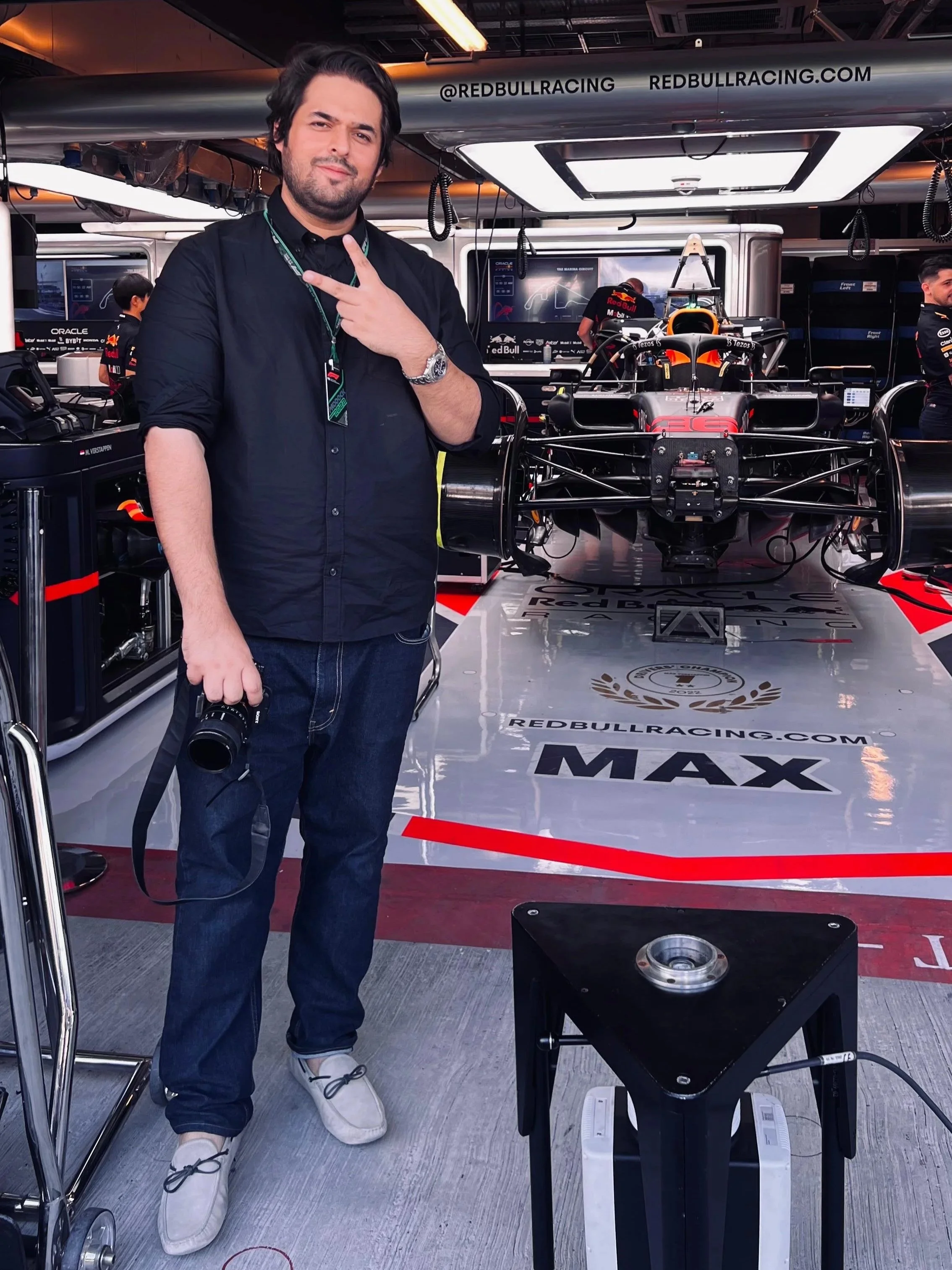 A man standing in a garage or workshop, holding a camera, with a Formula 1 race car behind him on a raised platform. The garage has Red Bull Racing branding and equipment, and other team personnel are working in the background. this is zain Jafar