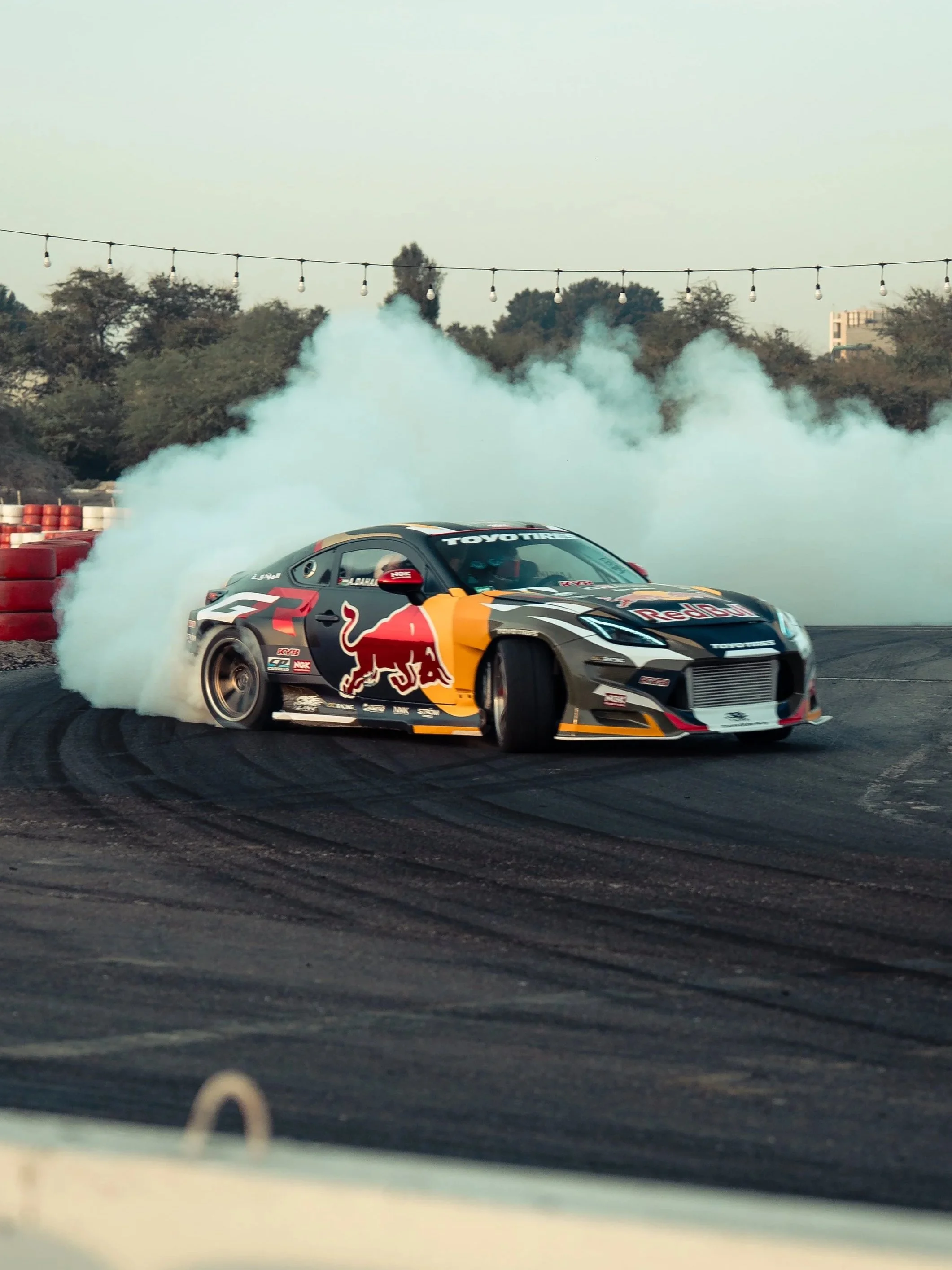 A black and red race car with Red Bull logo, drifting on a race track with smoke coming from its tires, surrounded by red and white barriers, with trees and a building in the background. ahmad daham drifting his drift car in jebel Ali