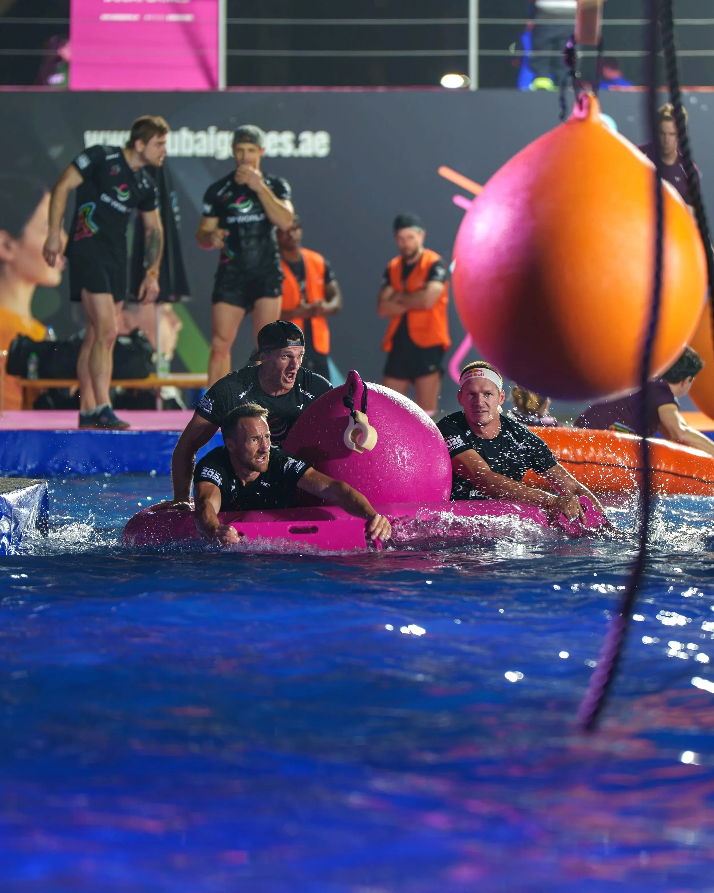 People in sports uniforms participating in a water event, with large orange inflatable balls, on a wet surface, possibly after a race or competition, with spectators or officials in the background. dubai games.