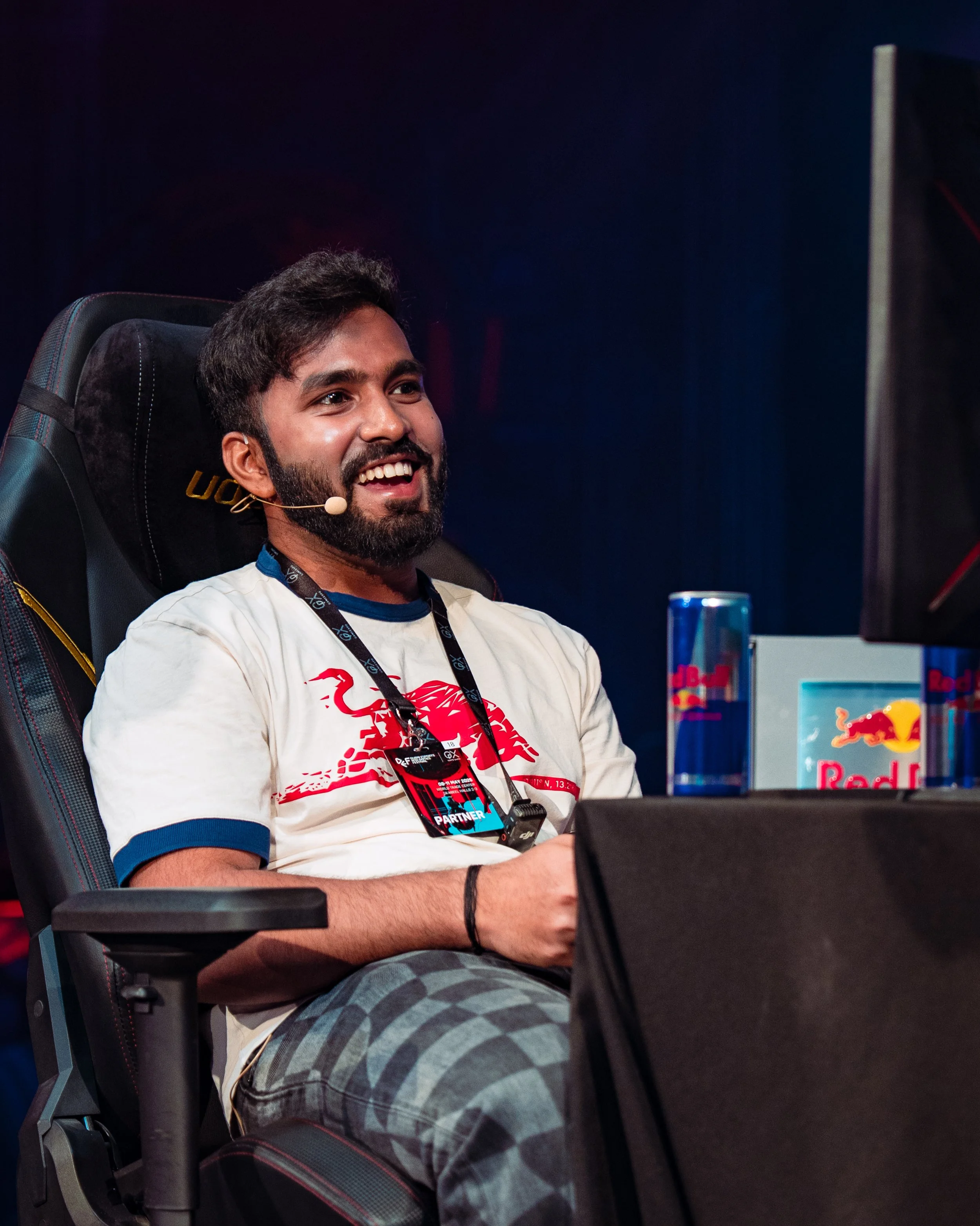 Smiling man with a beard and dark hair, wearing a white t-shirt with red graphics, sitting in a gaming chair with a headset microphone, looking at a computer monitor.