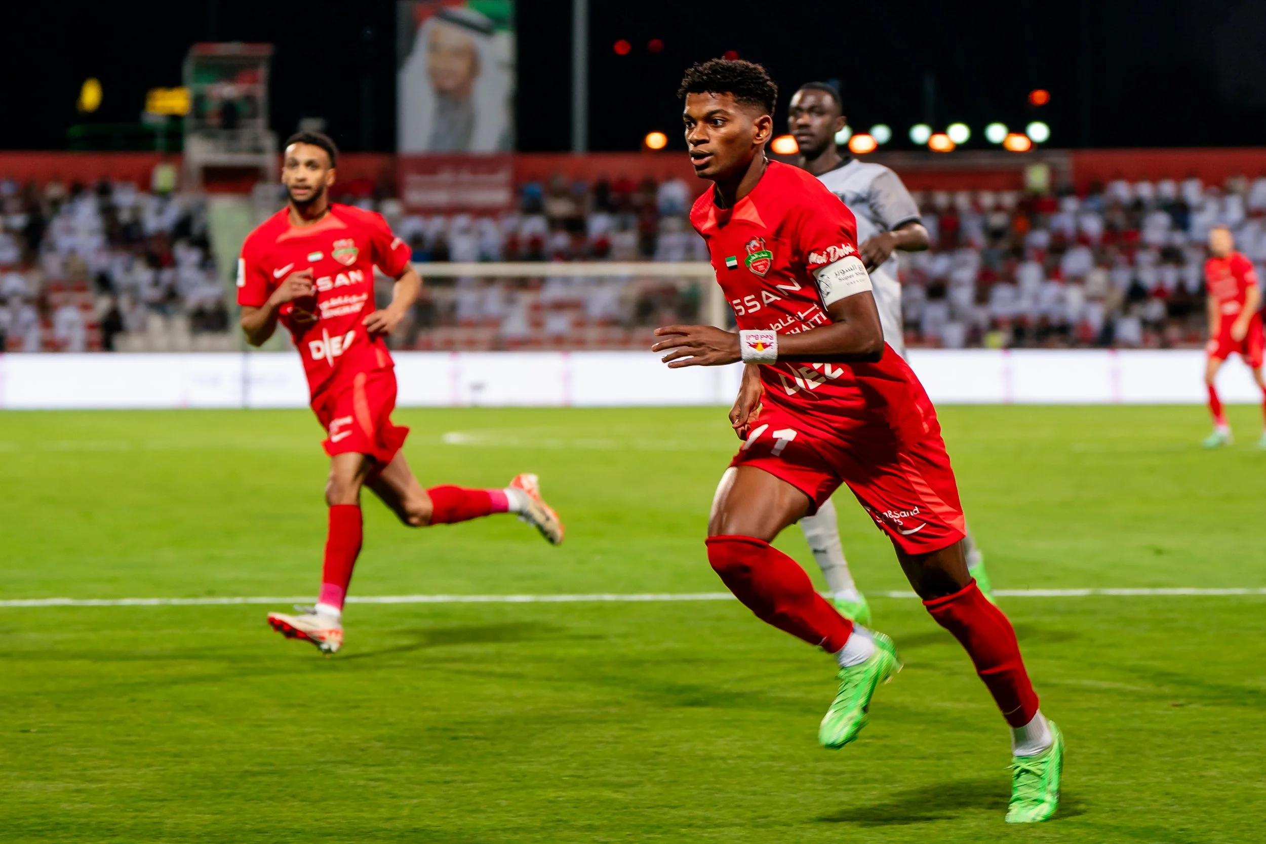 Soccer players in red uniforms playing on a green field at night, with a crowd in the stands and stadium lights in the background. Yahya Ghassan. the football player in the Uae pro league