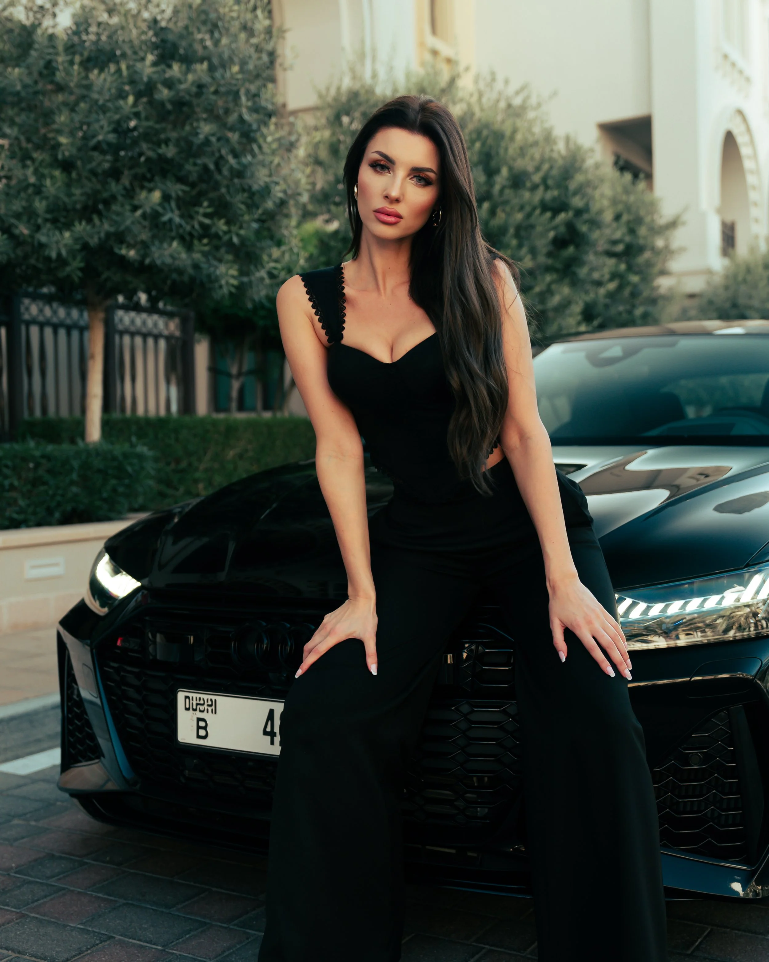 A woman with long dark hair, wearing a black sleeveless dress, posing with her hands on the front of a black luxury car. She is standing outdoors in front of a house with green trees in the background.