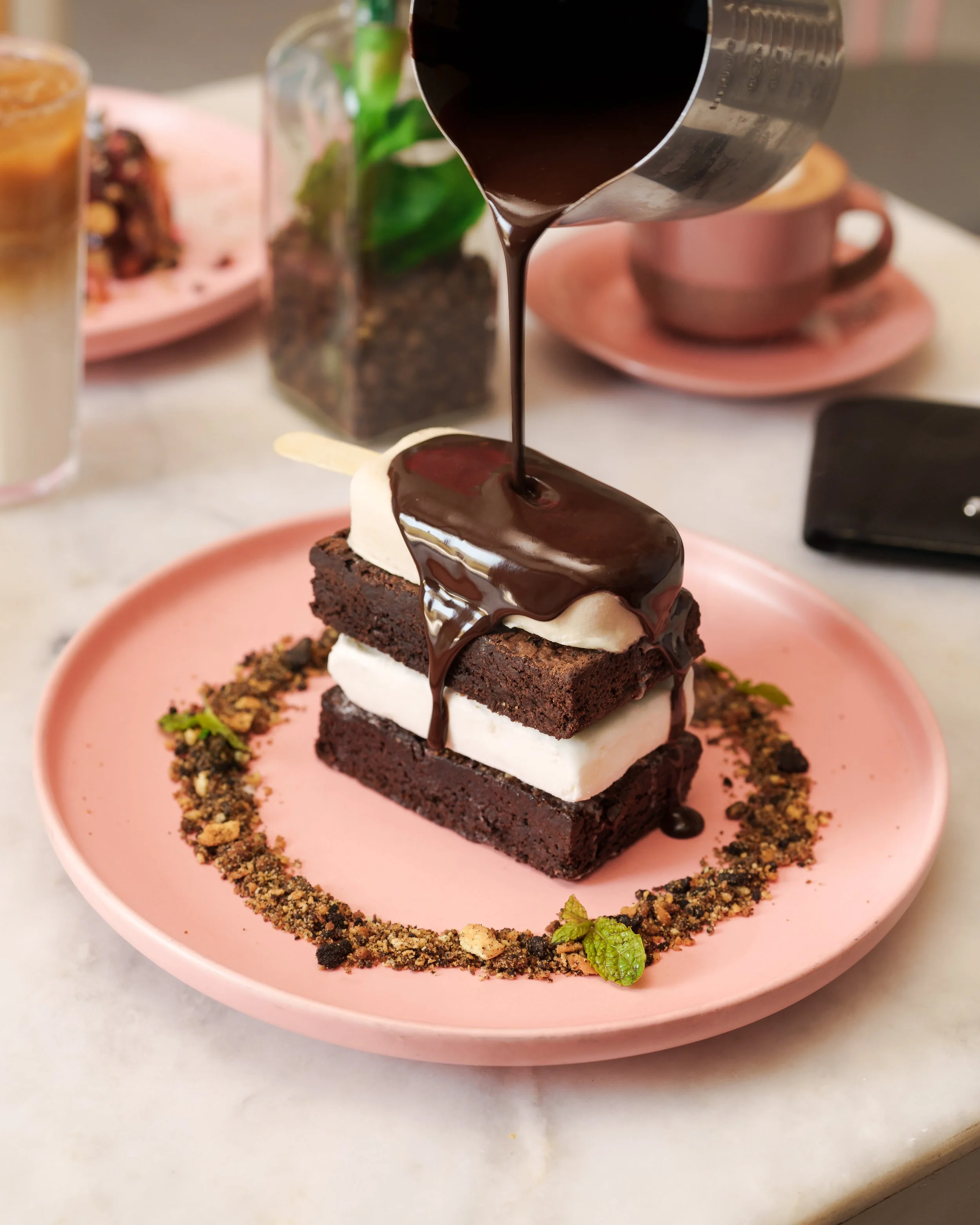 Chocolate dessert with white cream, topped with chocolate sauce and placed on a pink plate, garnished with crushed nuts and mint leaves, with coffee and a drink in the background. food photography in dubai. for caffe Praha