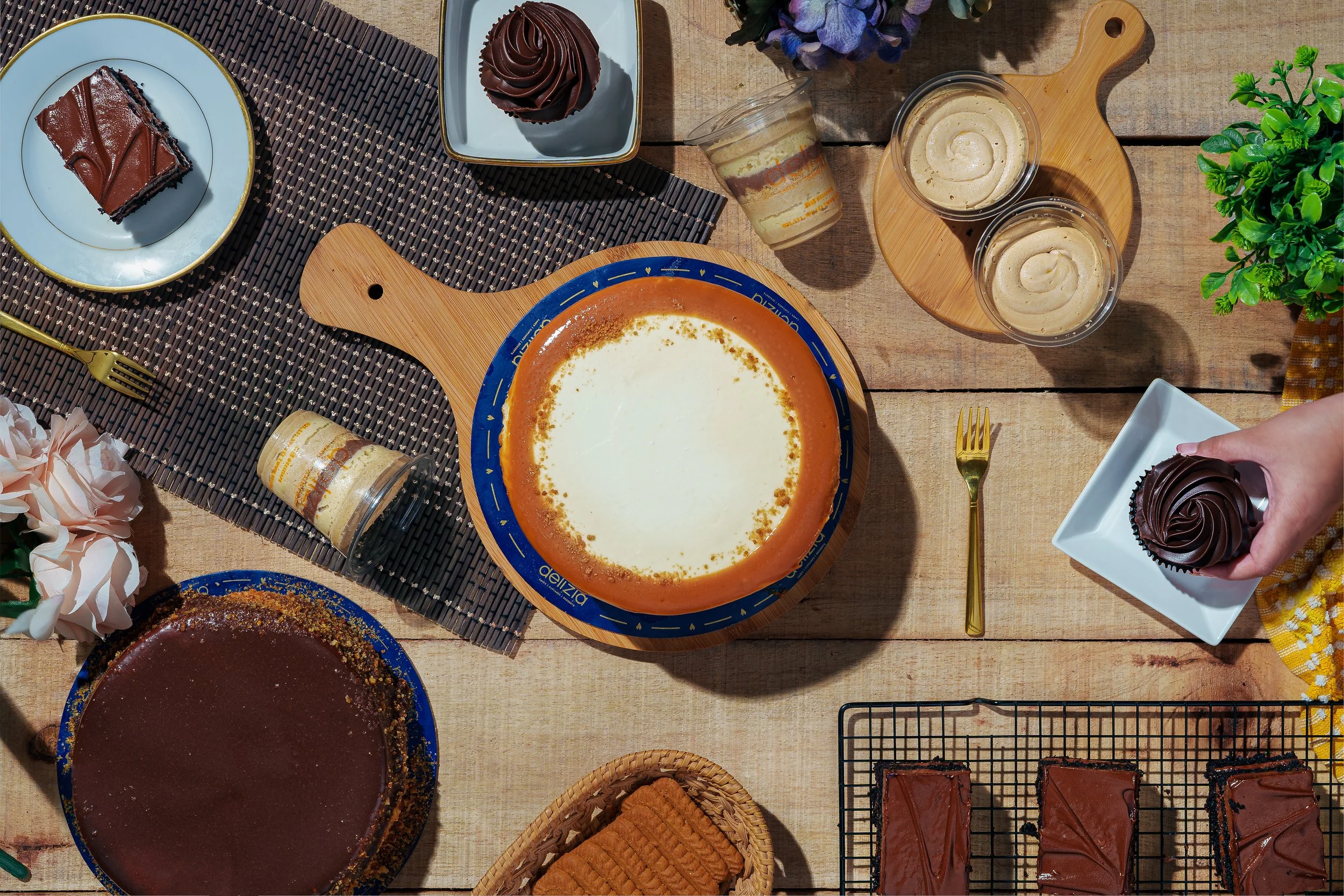 A top-down view of a dessert table with chocolate cake, cupcake, cookies, puddings, and flowers on a wooden table. Delizia. food photography in dubai