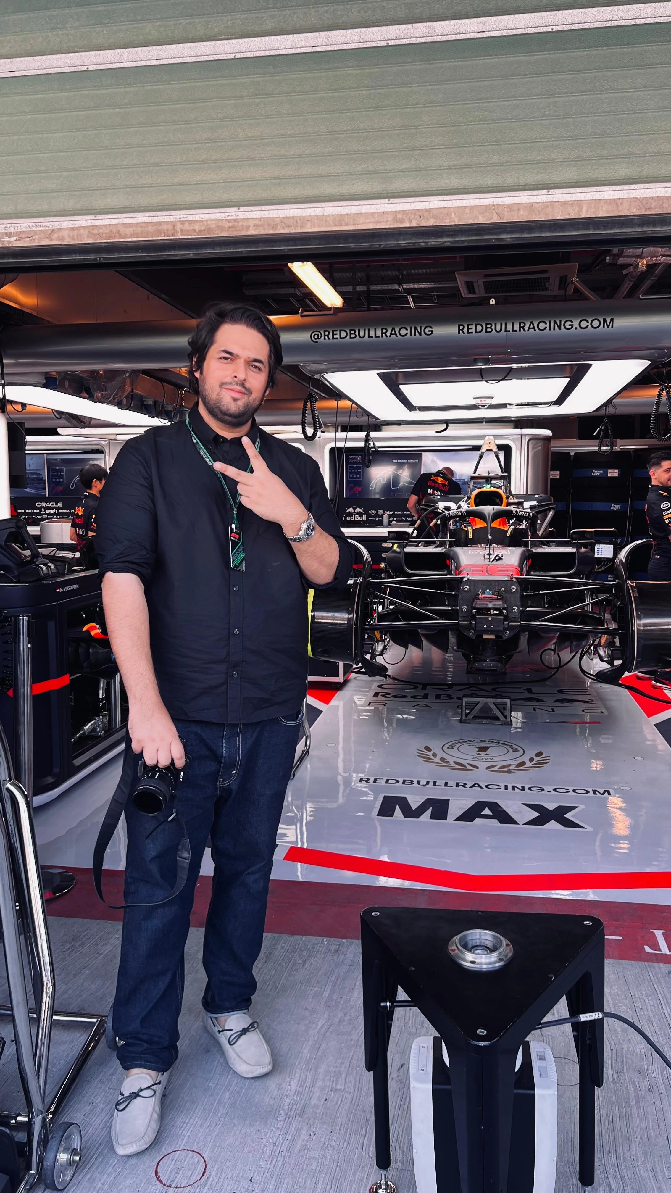 Man in black shirt and jeans standing in a racing garage with a racing car behind him. The garage has Red Bull Racing branding, tools, and equipment displayed.