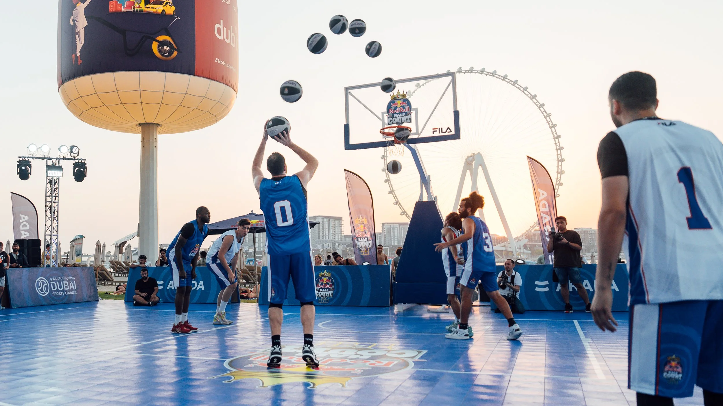 A basketball game taking place outdoors on a city rooftop, with players in blue jerseys, some holding basketballs, and a Ferris wheel in the background at sunset. sequence photography.