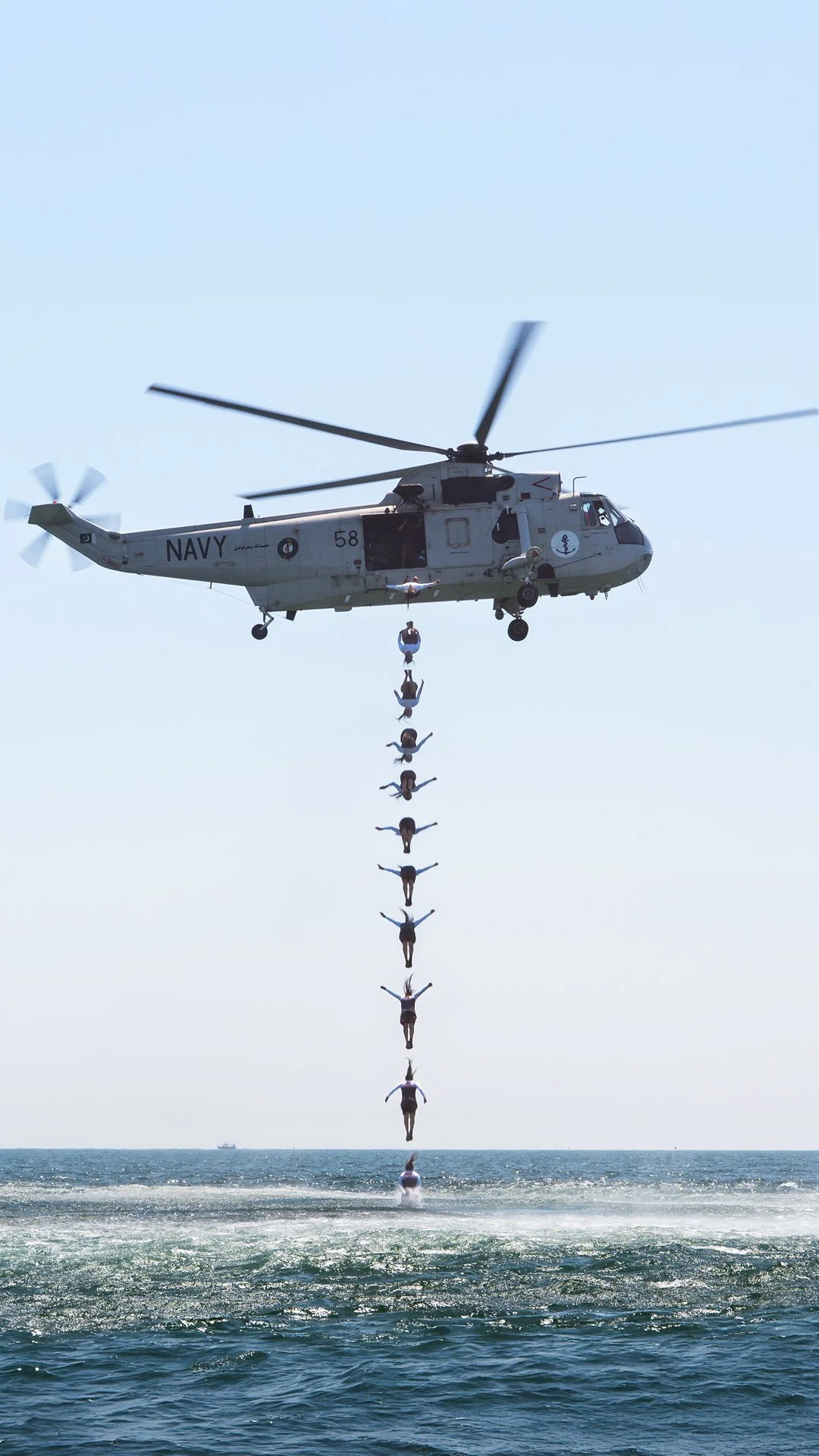 Several people are performing a synchronized diving routine from a helicopter over the water, forming a vertical line with their bodies extended outward. just one person. 
sequence shot of Orlando duque jumping out of a helicopter in pakistan.