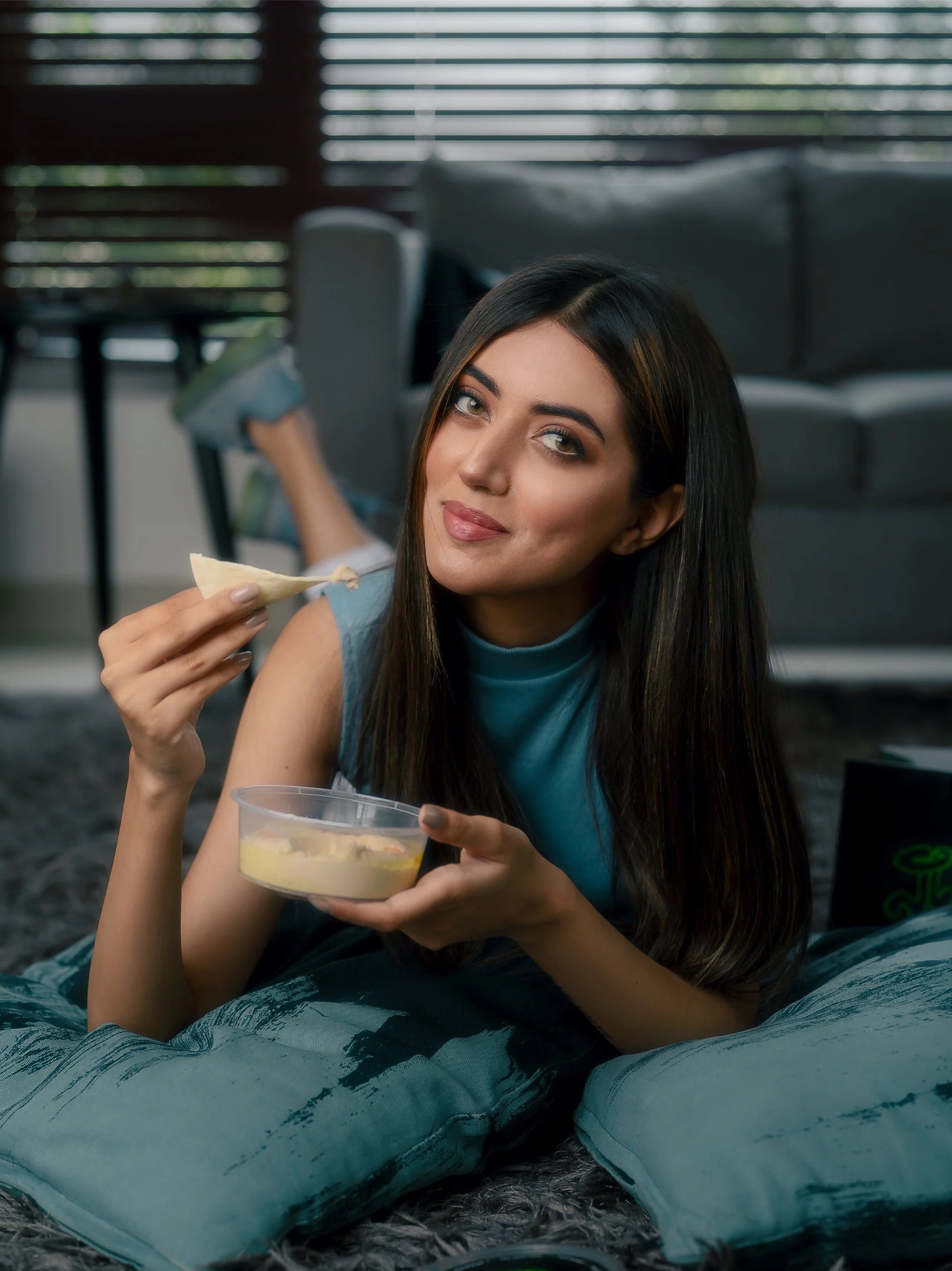 A woman with long dark hair lying on a bed with patterned blankets, holding a small bowl of dip or sauce and a tortilla chip, smiling at the camera.
