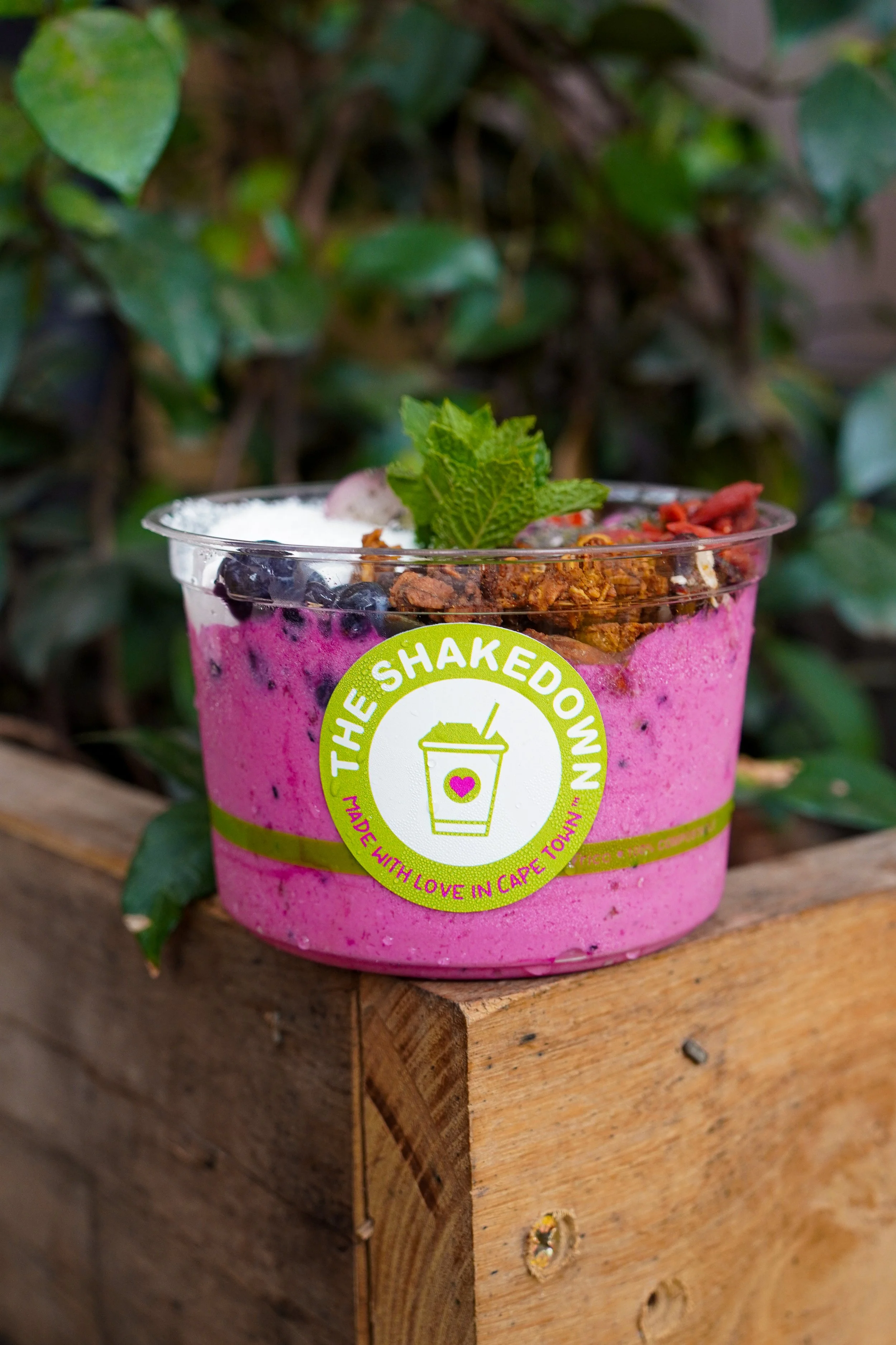 A Dragon Fruit smoothie in a clear cup with blueberries, granola, mint leaves, and other toppings, placed on a wooden surface outdoors.