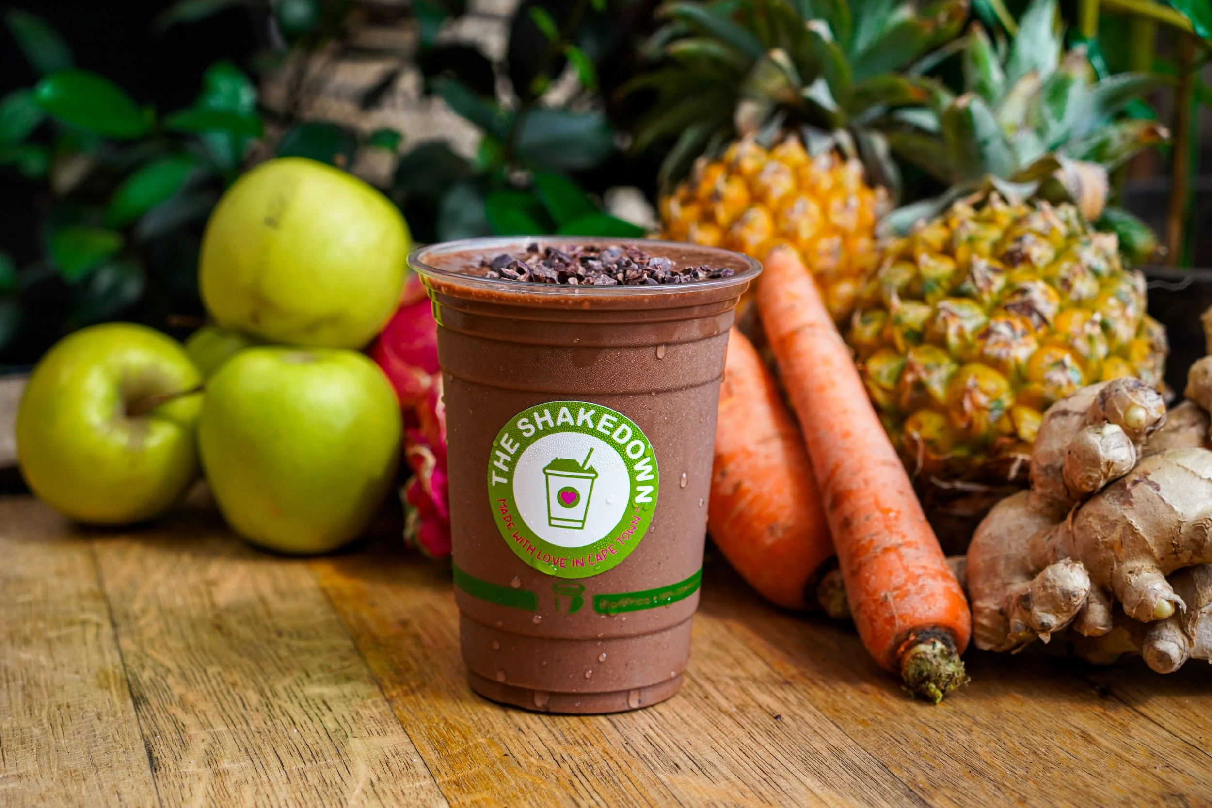 A chocolate smoothie in a cup with the logo of The Shakedown, surrounded by fresh green apples, carrots, pineapples, and ginger on a wooden surface.