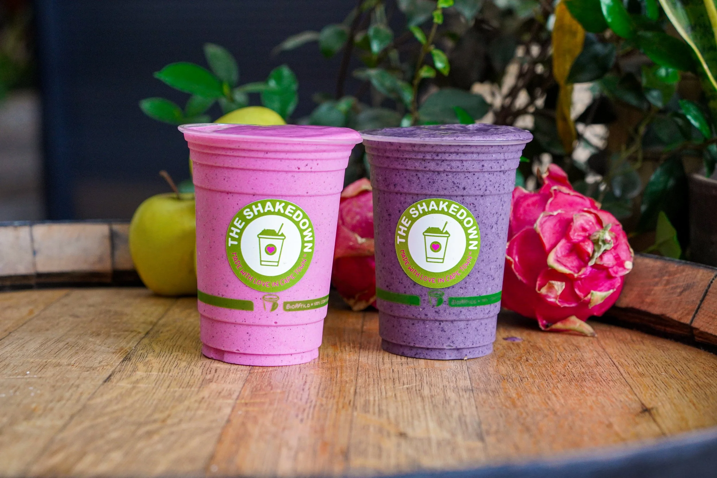 Two colorful smoothies in plastic cups with the logo 'The Shakedown' on a wooden table, with green apples and pink dragon fruit in the background.