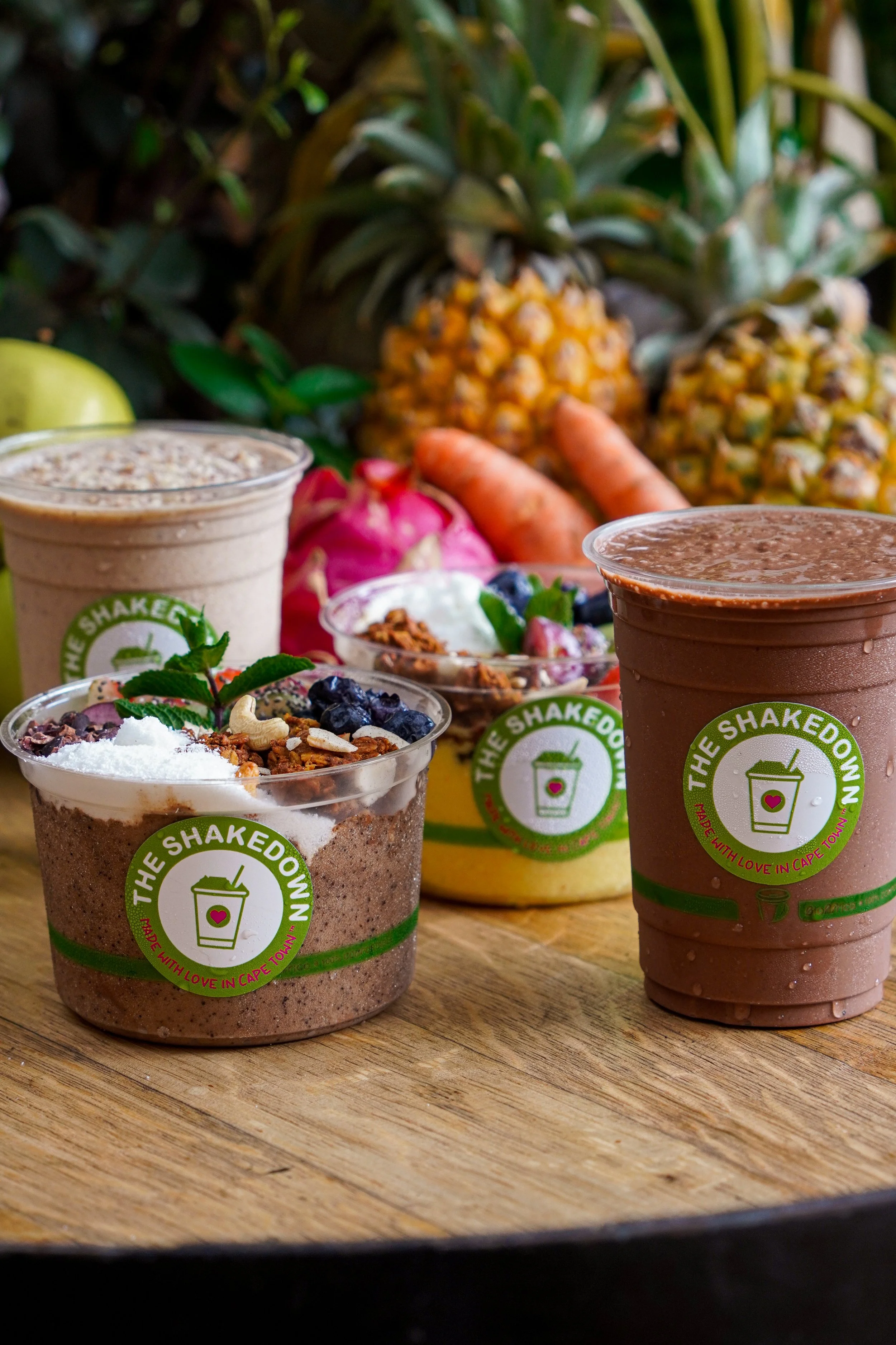 Various fruit smoothies and bowls with fruits, carrots, pineapples, apples, and herbs on a wooden table.