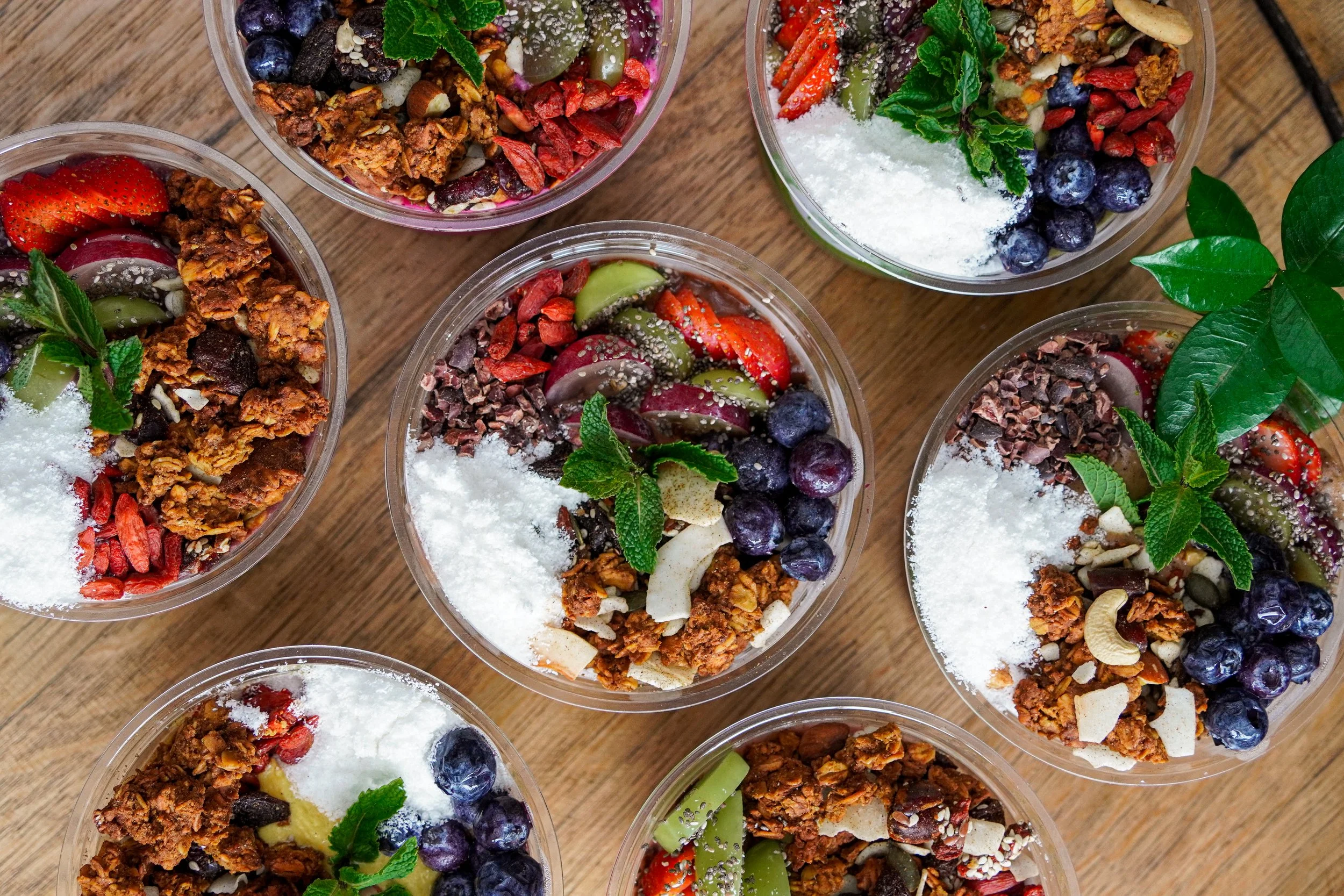 Multiple clear plastic bowls with various fruit and granola parfaits on a wooden surface, topped with fresh berries, mint leaves, and shredded coconut.