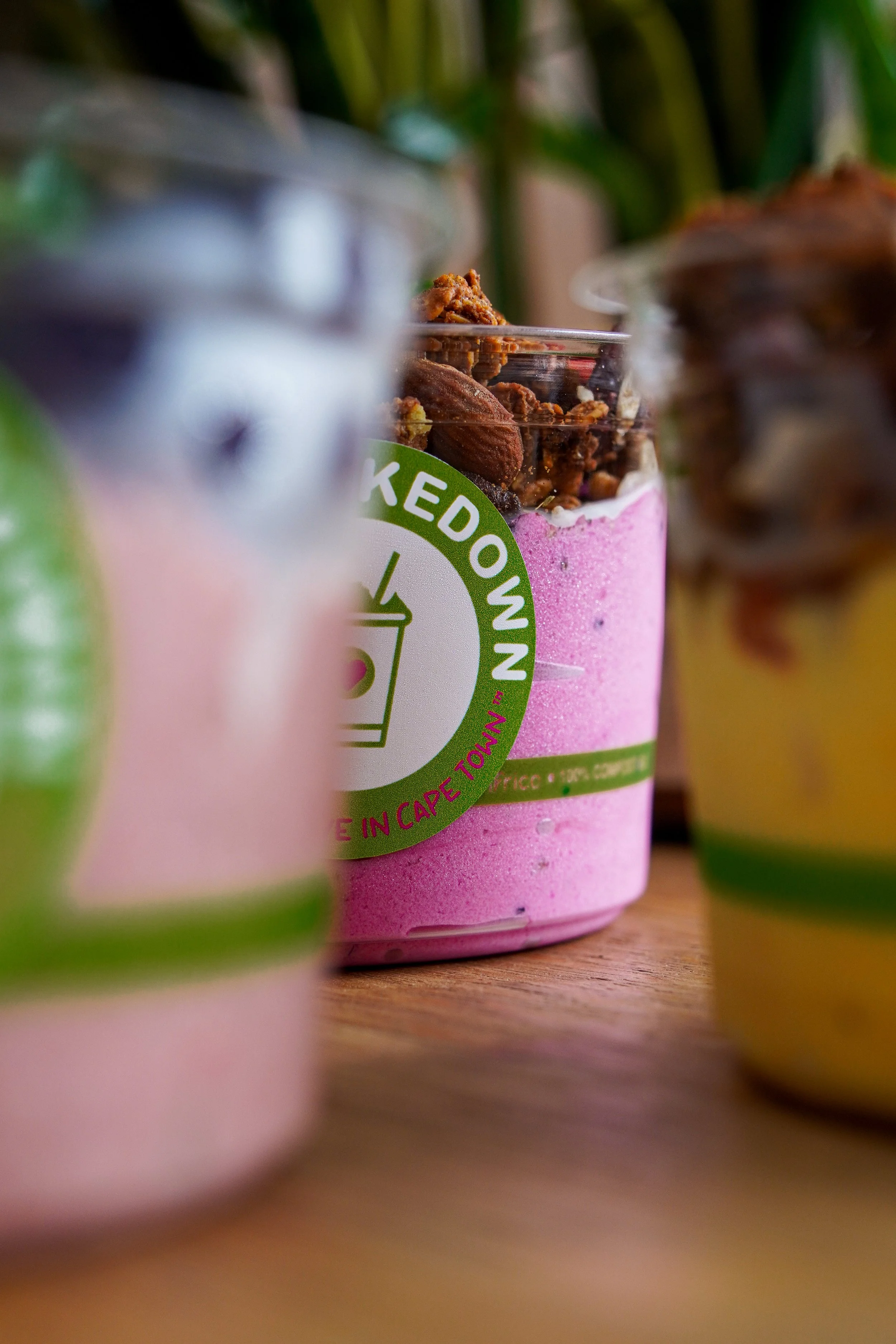Close-up of a pink smoothie in a clear cup with a green and white logo, topped with granola, oats, and nuts, with blurred green and yellow objects in the foreground and background.