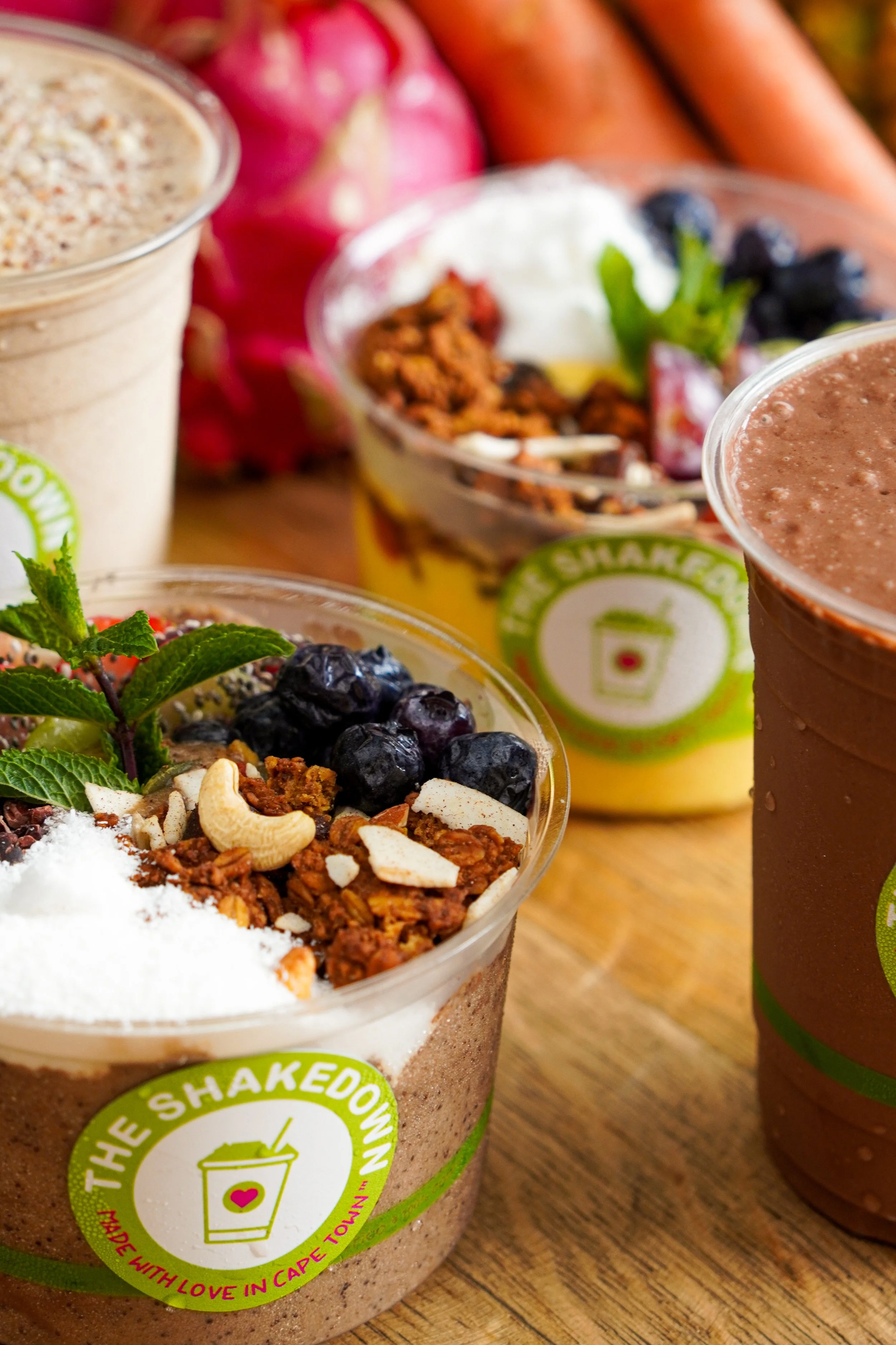 Close-up of a fruit and granola yogurt parfait with blueberries, coconut flakes, and mint, next to a chocolate smoothie and a vanilla smoothie, on a wooden surface.