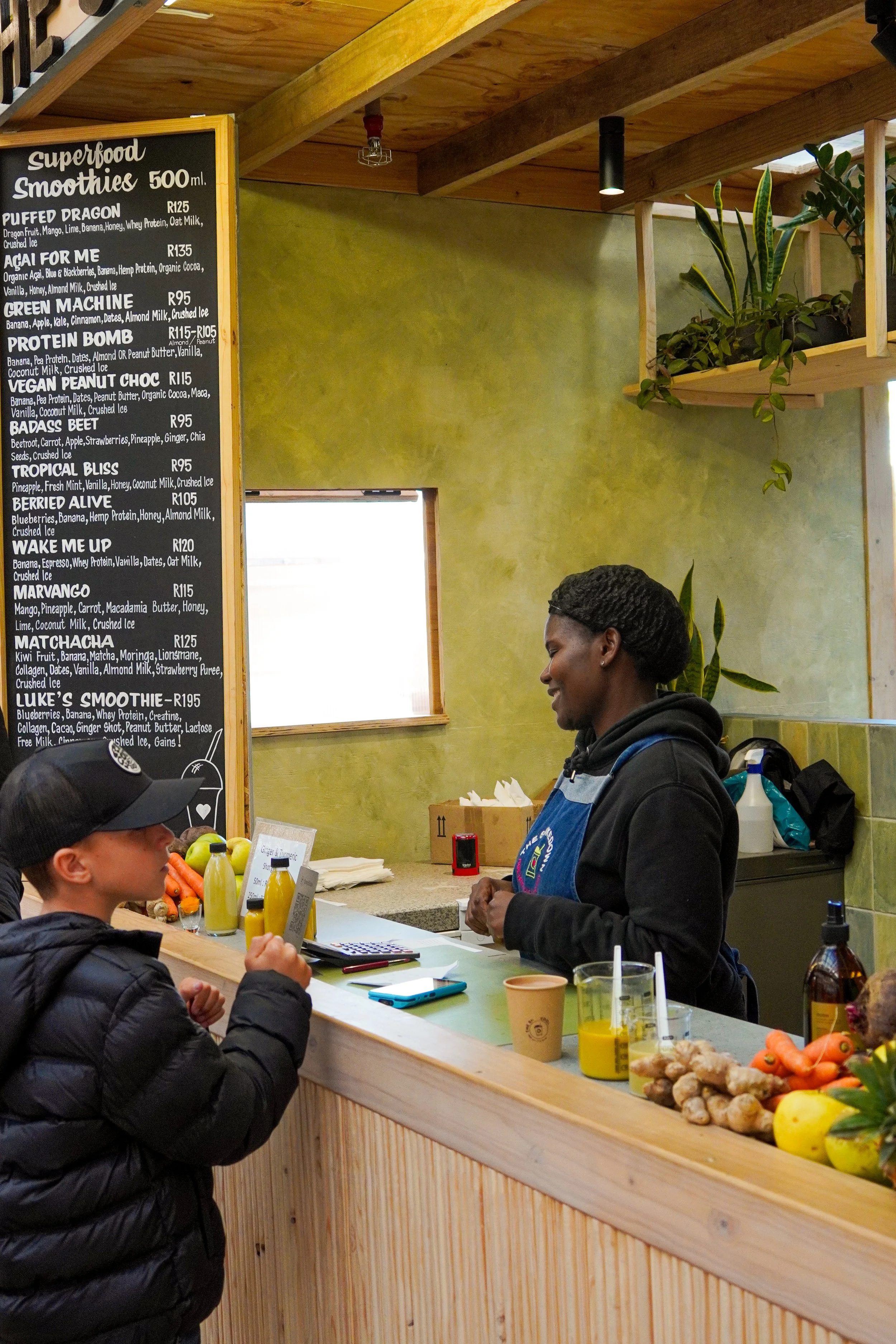 A woman in a black hoodie and an apron working at a counter inside a green-walled shop; she is talking to a young boy wearing a black jacket and hat. The counter has various fruits and vegetables, juice bottles, and paper cups. A blackboard menu displays smoothie options with prices.