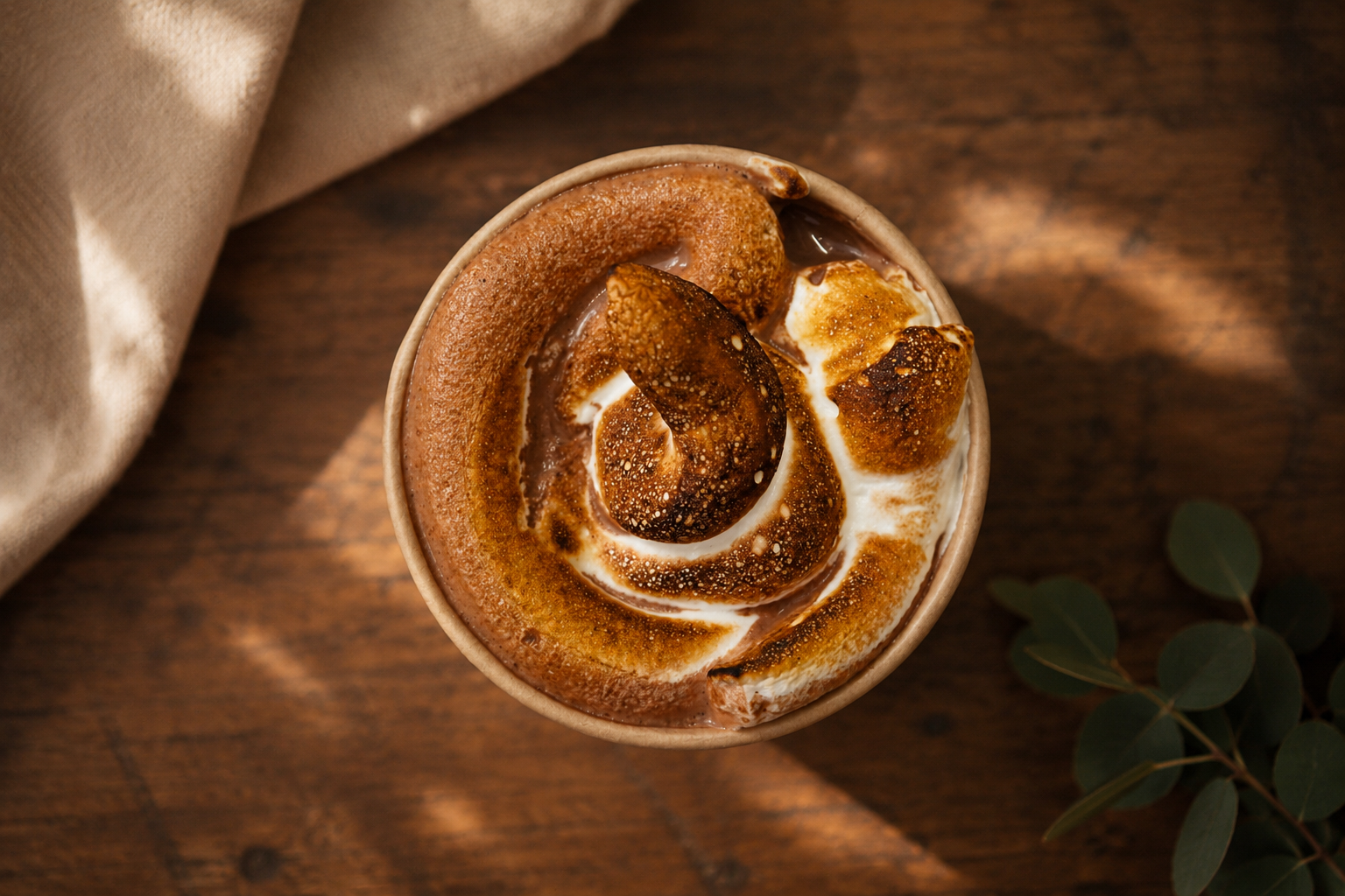 Coffee drink topped with whipped cream, cinnamon, and caramelized sugar, placed on a wooden table with a cloth and eucalyptus leaves nearby.