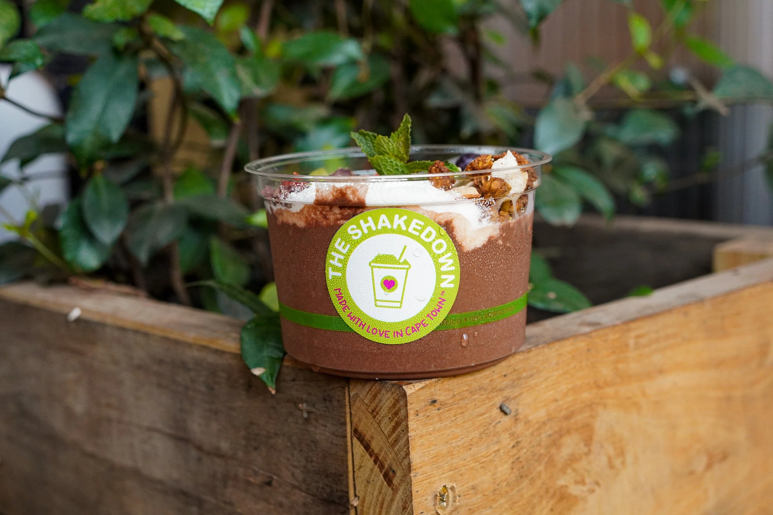 A plastic cup with a green and white label reading 'The Shakedown' and 'Made with Love in Cape Town' contains a layered dessert topped with whipped cream, granola, a sprig of mint, and cocoa powder, set on a wooden surface outdoors with green leafy plants in the background.