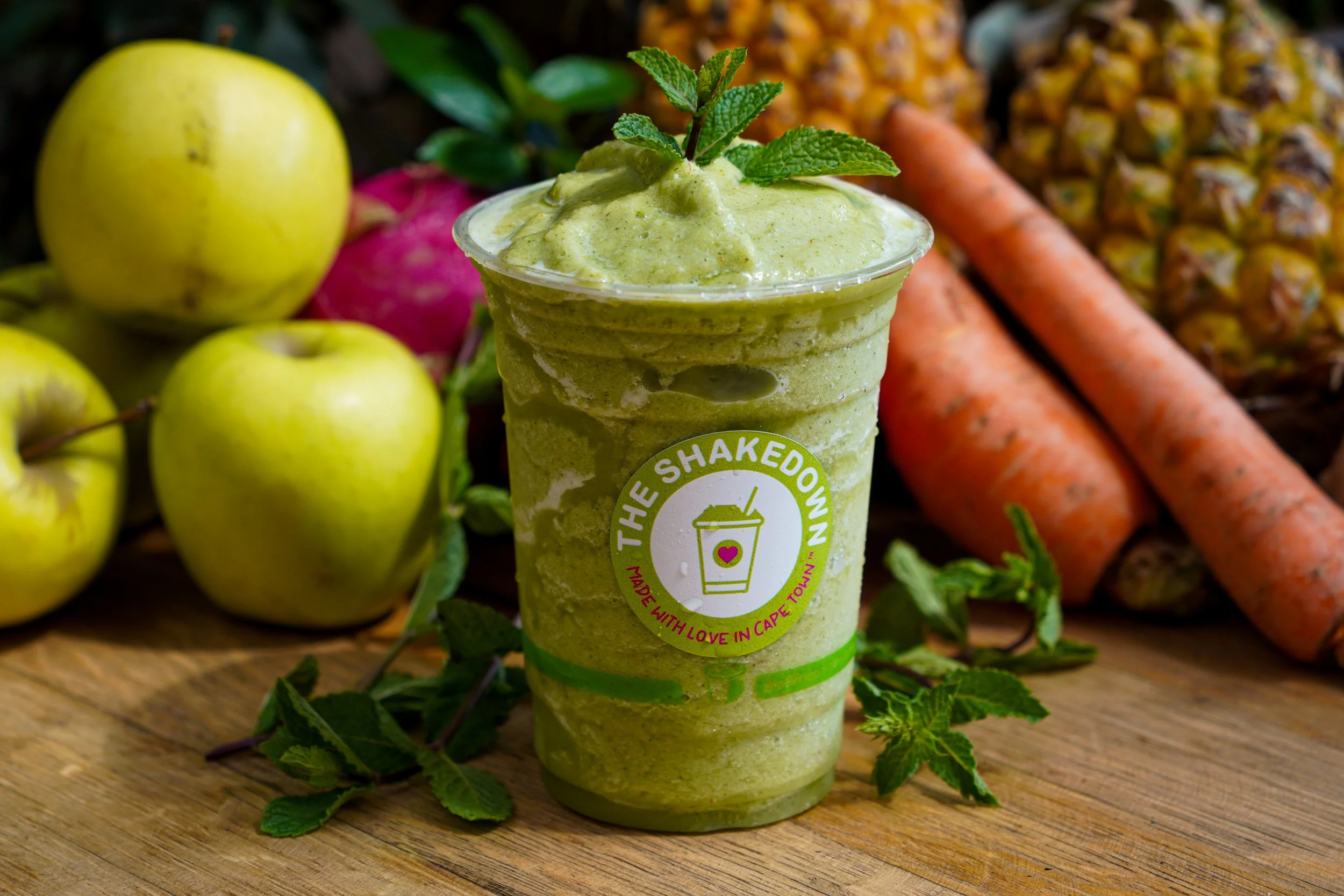 Green smoothie in a clear cup garnished with mint leaves, surrounded by green apples, carrots, pineapple, and mint leaves on a wooden surface.