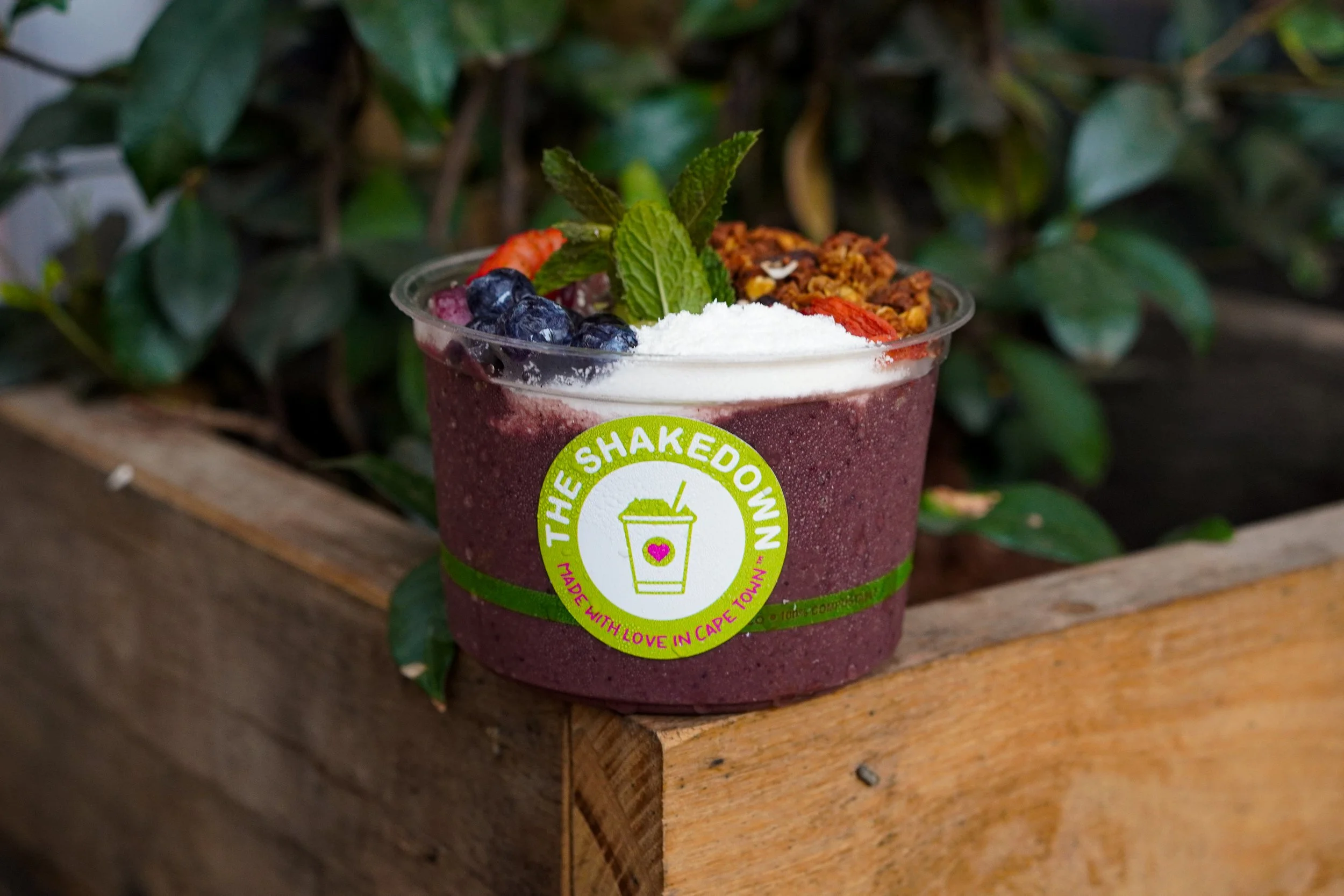 A fruit smoothie in a clear cup topped with blueberries, goji berries, granola, a mint sprig, and a scoop of white powder. The cup has a green and pink label from The Shakedown with the text "Made with love in Cape Town." The cup is placed on a wooden surface in front of green leafy plants.