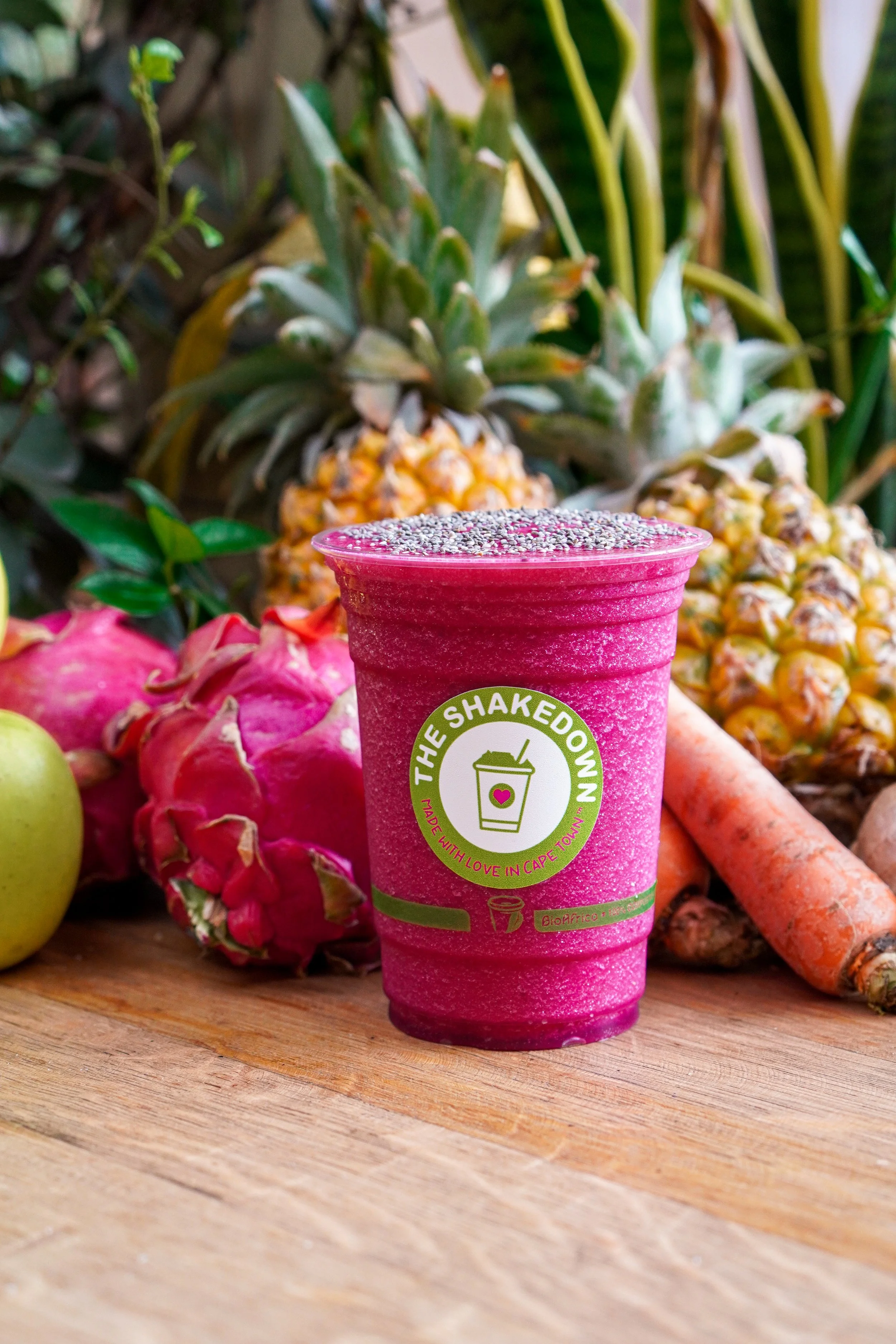 Pink smoothie cup with label in front of tropical fruits including pineapples, dragon fruit, and carrots on a wooden surface.