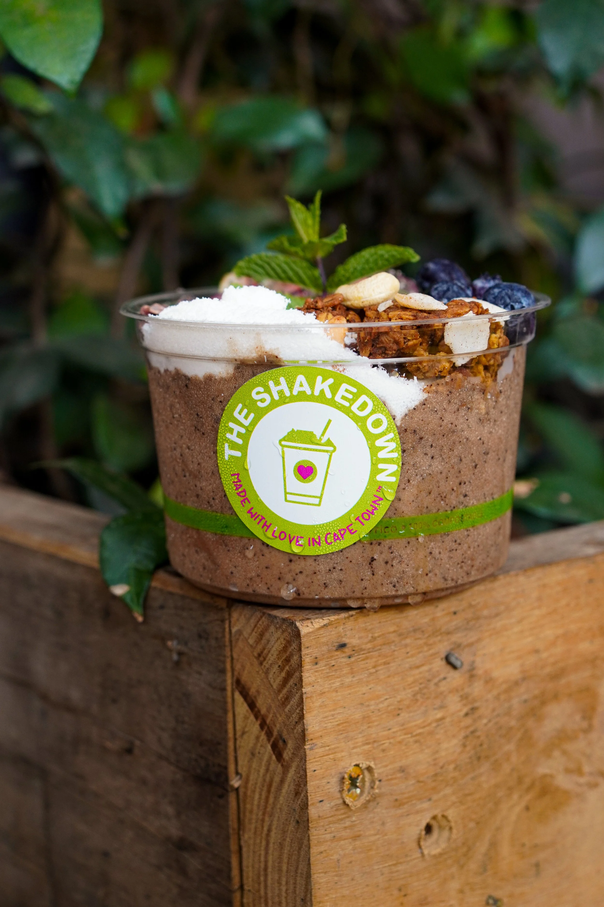 A healthy smoothie bowl in a clear cup with a green label, topped with granola, nuts, dried blueberries, and fresh mint, placed on a wooden surface with green foliage in the background.