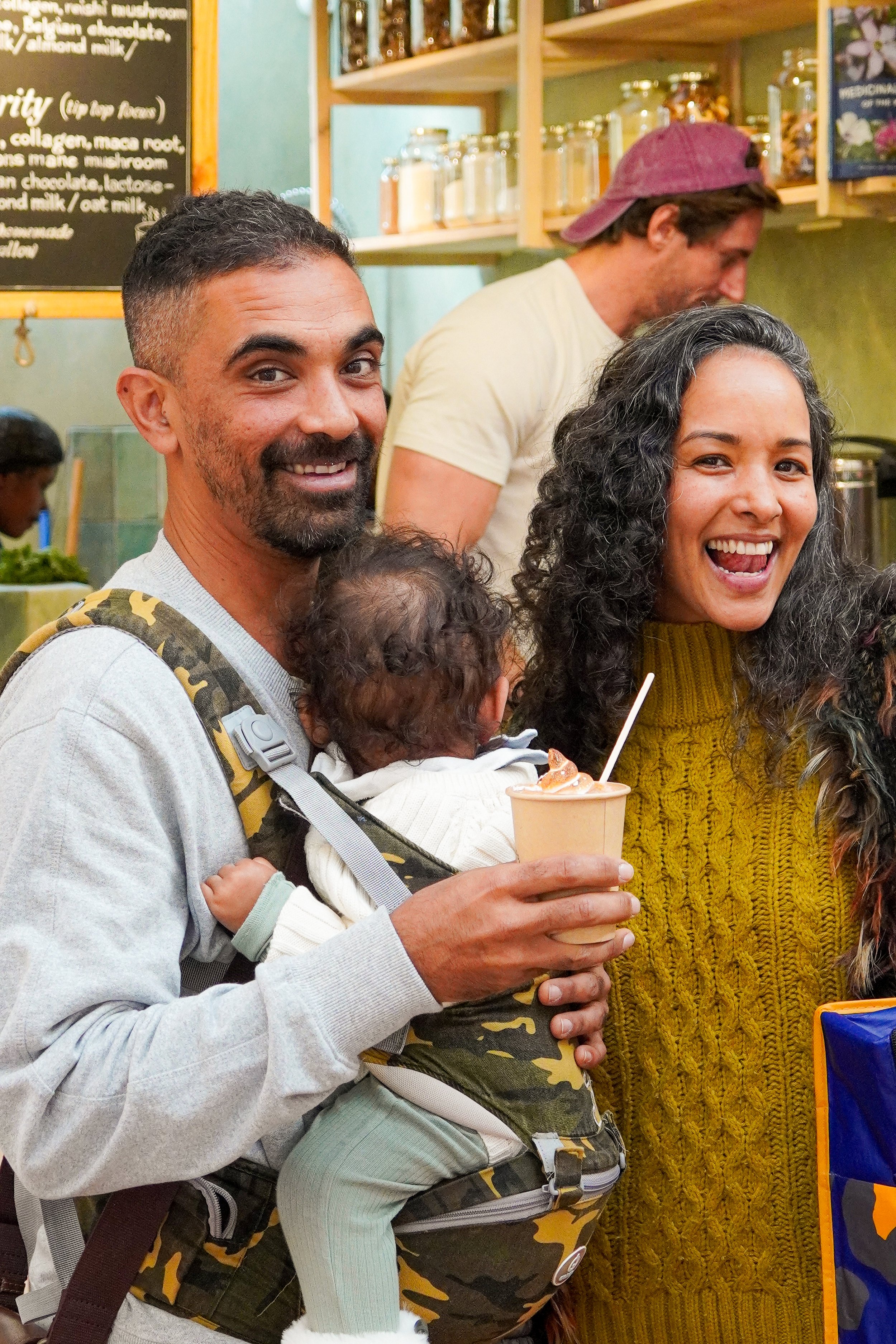 A happy man with a beard and a woman smiling, both holding a young child in a baby carrier. The man is holding a cup of coffee. They are in a cafe with shelves of jars in the background.