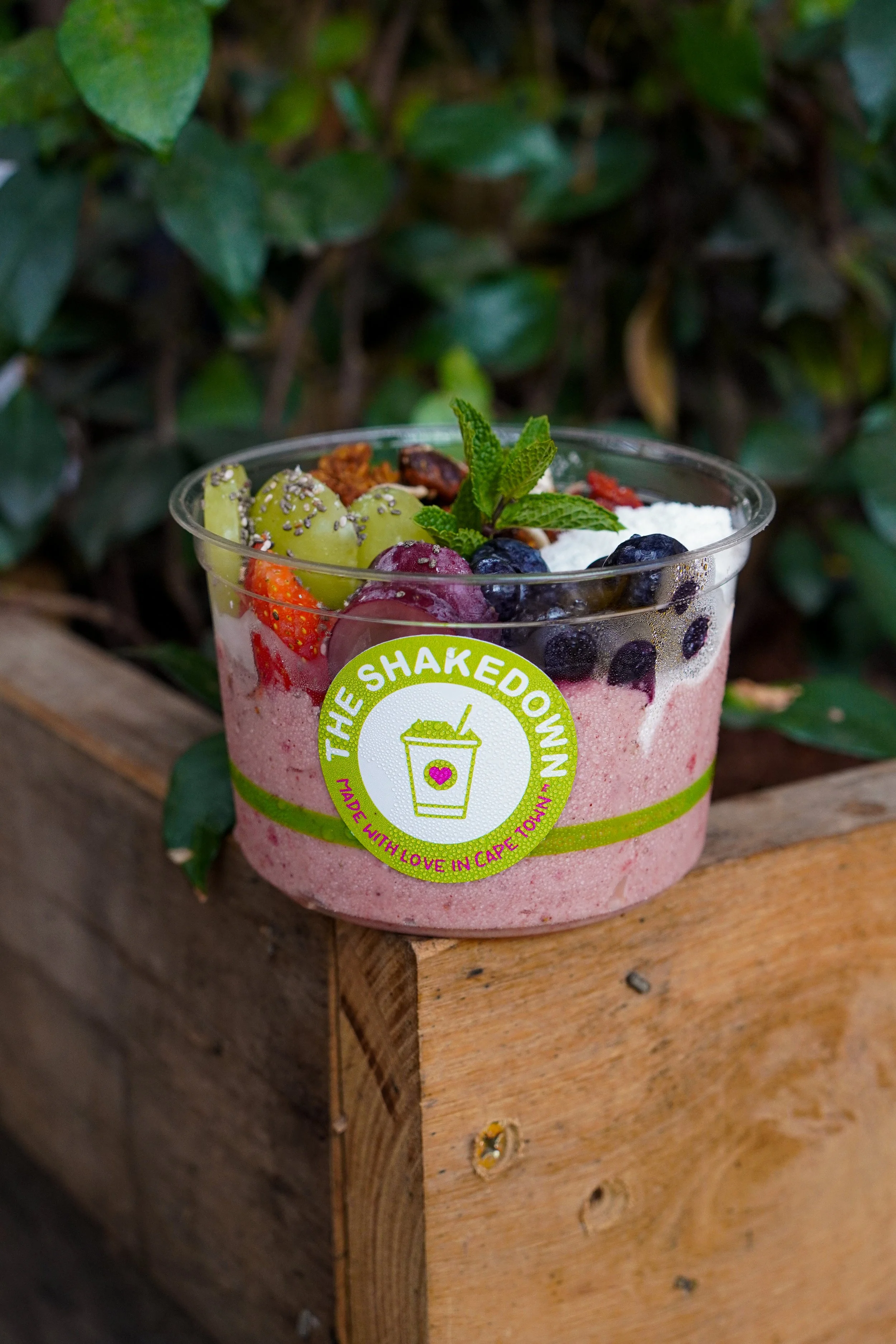 Close-up of a pink smoothie in a clear cup with a lime green sticker, topped with strawberries, blueberries, grapes, nuts, and mint leaves, placed on a wooden surface with green foliage in the background.
