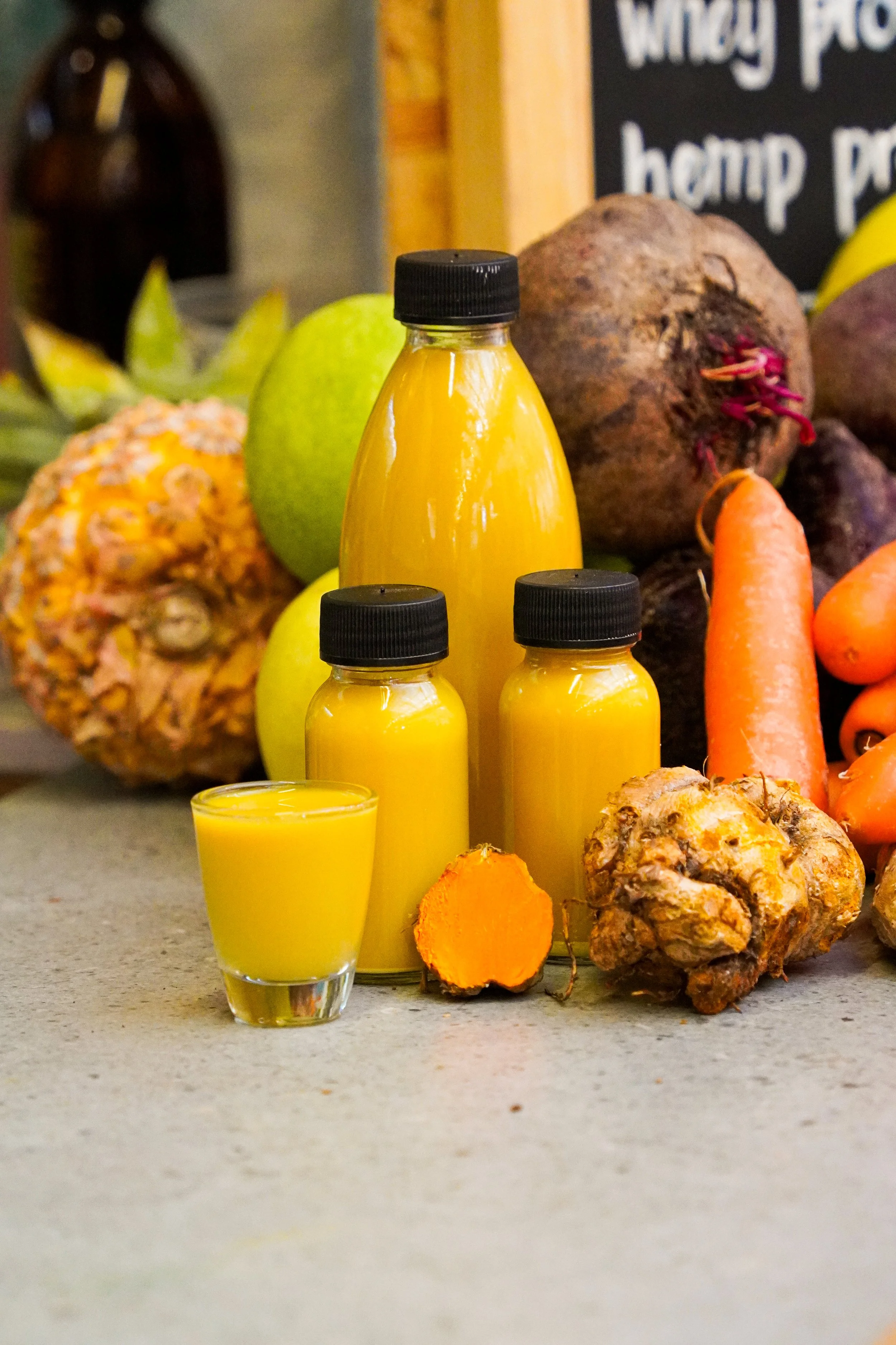 A variety of tropical fruits and vegetable juices on a countertop, including pineapples, a green melon, beets, carrots, and ginger root, with bottles of yellow juice and a glass of fresh juice in front.