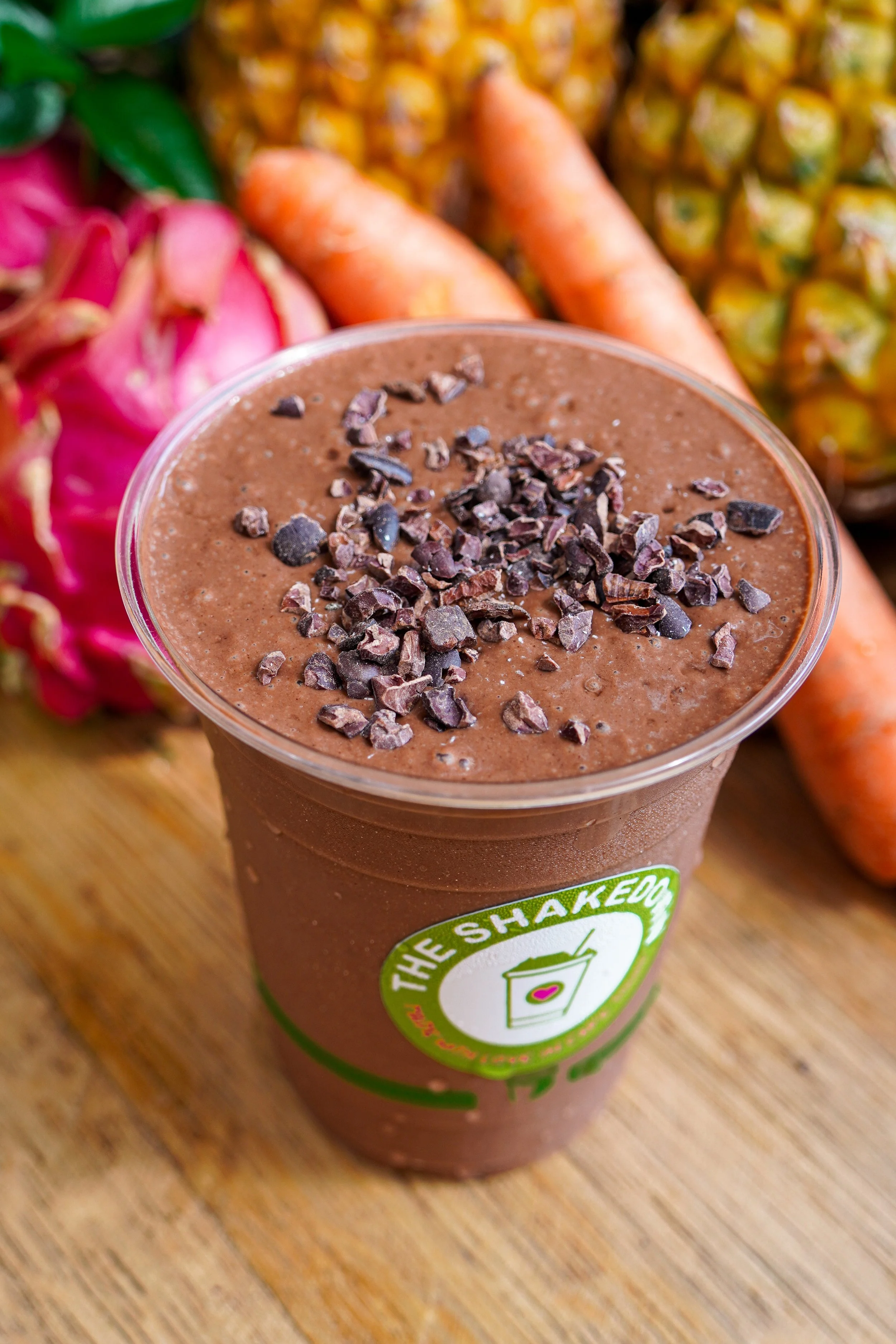 A chocolate smoothie in a clear cup topped with chocolate shavings, placed on a wooden surface with fresh pineapples, carrots, and dragon fruit in the background.