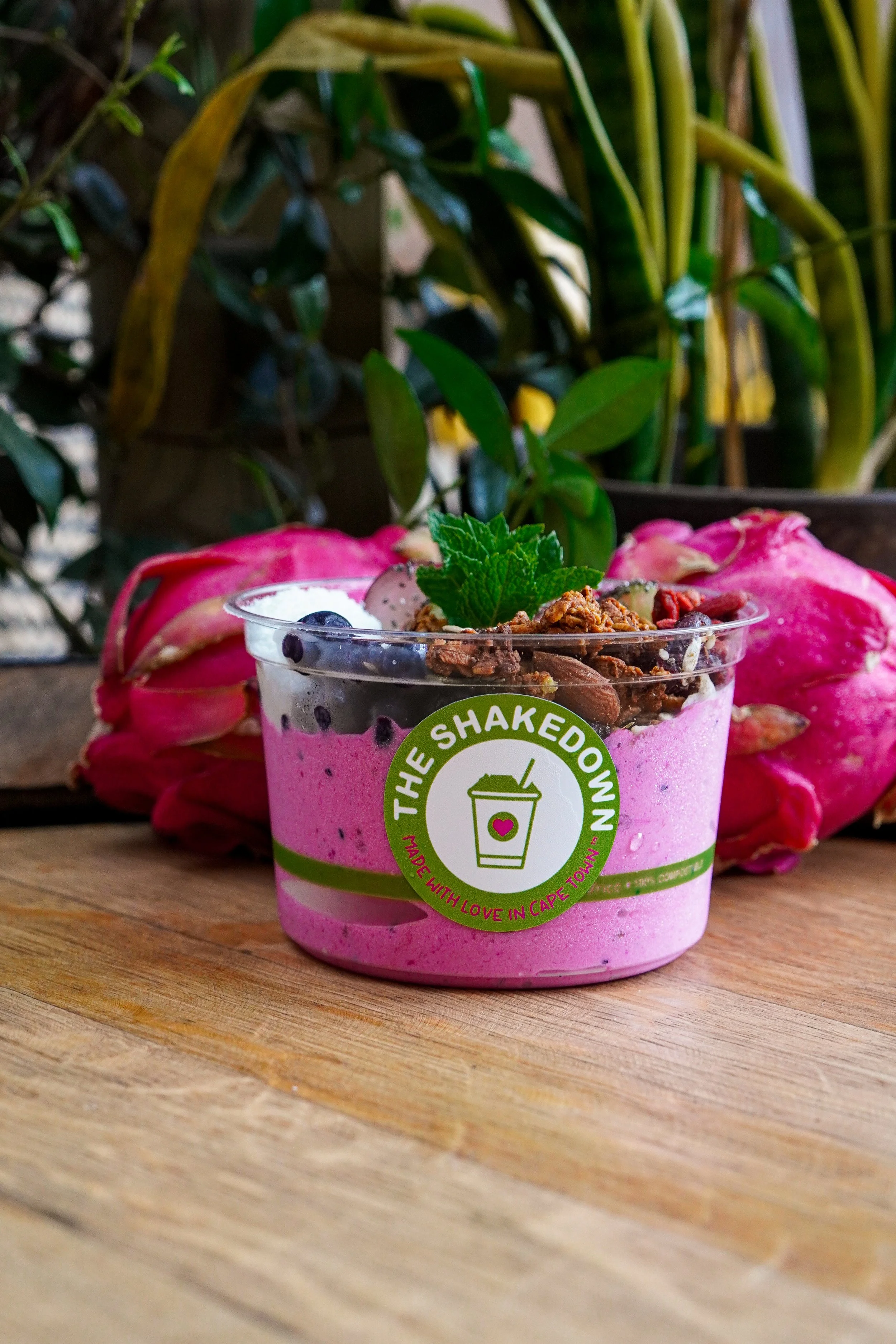 A pink smoothie cup with a green label reading "The Shakedown" and inner text "Made with love in Cape Town." The smoothie is topped with granola, blueberries, and a mint leaf, and is placed on a wooden surface backdrop of leafy green plants and pink dragon fruit.