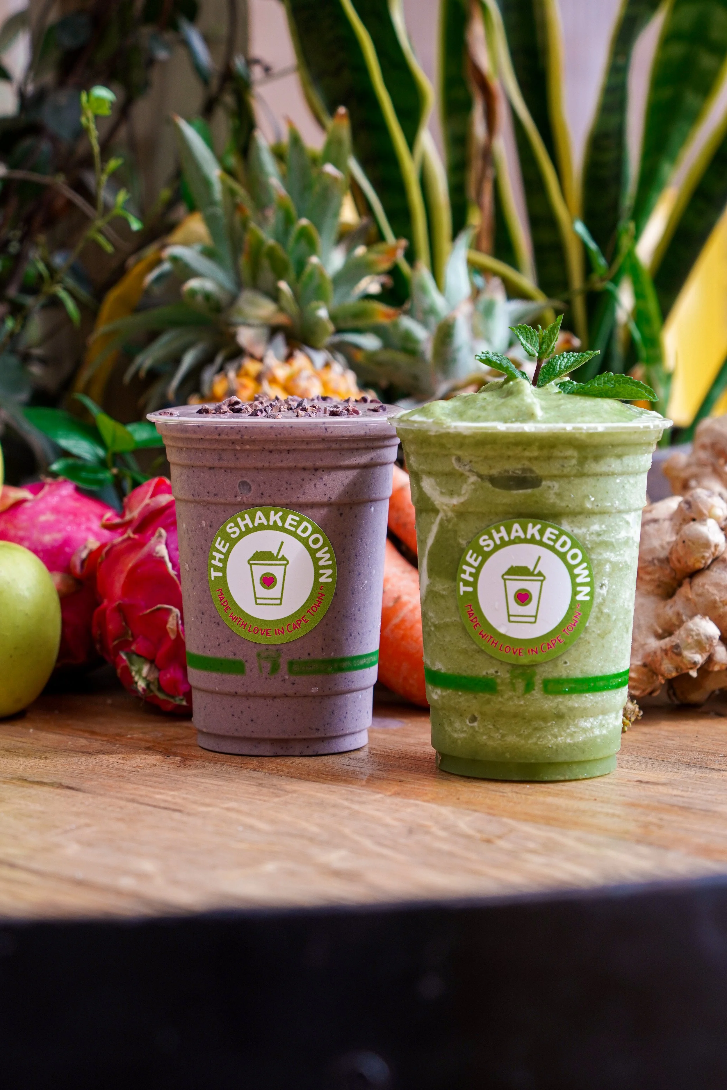 Two green smoothies in plastic cups with white labels that read 'The Shakedown' and 'Made with Love in Cape Town,' placed on a wooden surface. Behind the smoothies, there are various fruits including pineapples, apples, dragon fruits, and ginger, with green plants in the background.