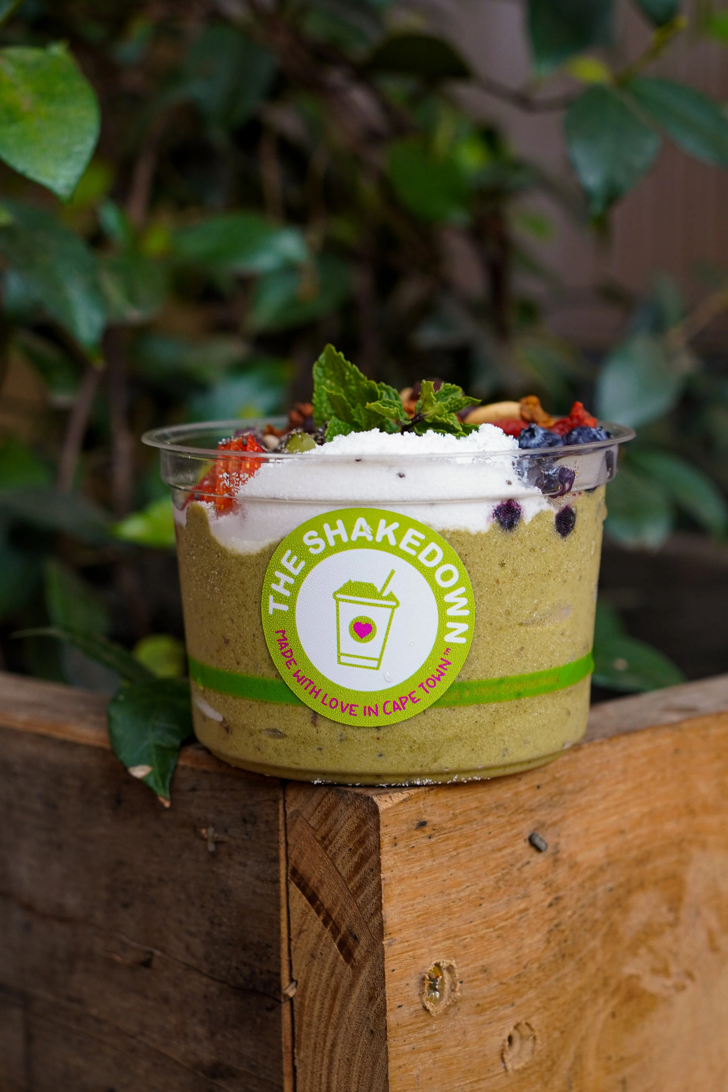 A green smoothie in a clear plastic cup with a label reading "The Shakedown" and the phrase "Made with love in Cape Town". The smoothie is topped with white powder, fresh berries, and mint leaves, placed on a wooden surface outdoors with green leaves in the background.