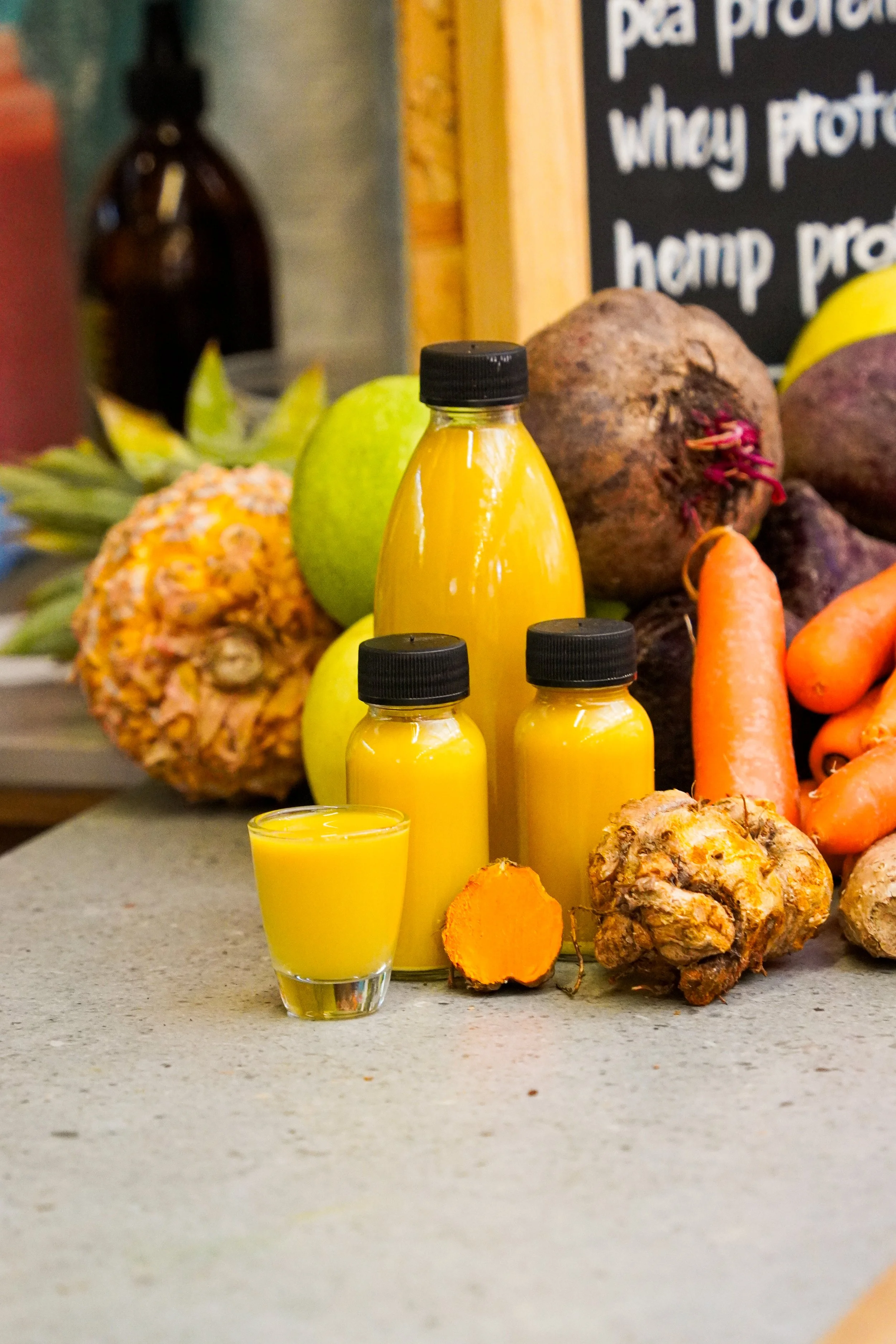 A display of fresh vegetables and bottles of yellow juice on a countertop, with a handwritten menu board in the background.