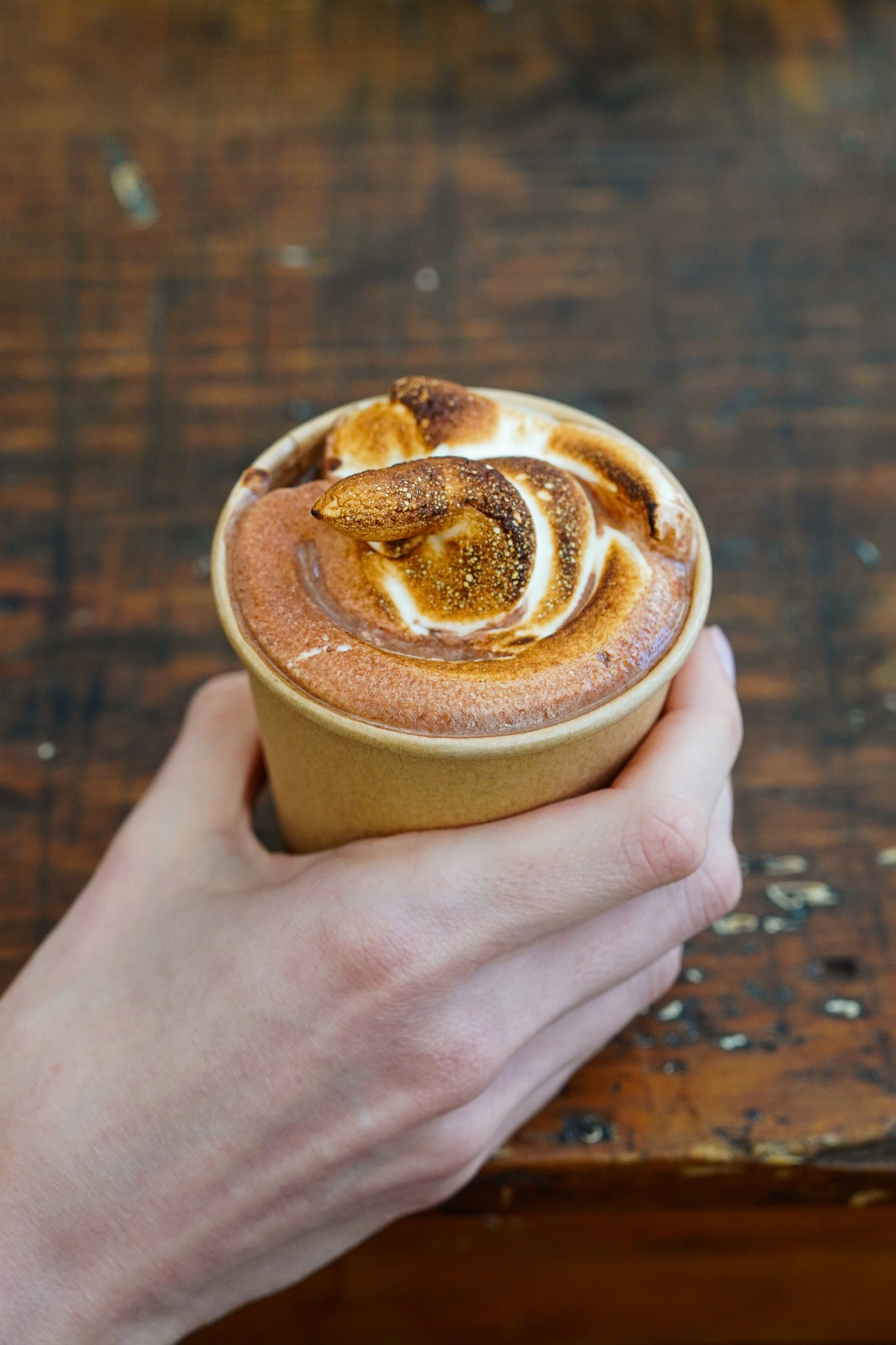A hand holding a cup of coffee topped with whipped cream and sprinkled with cinnamon, set against a wooden table background.