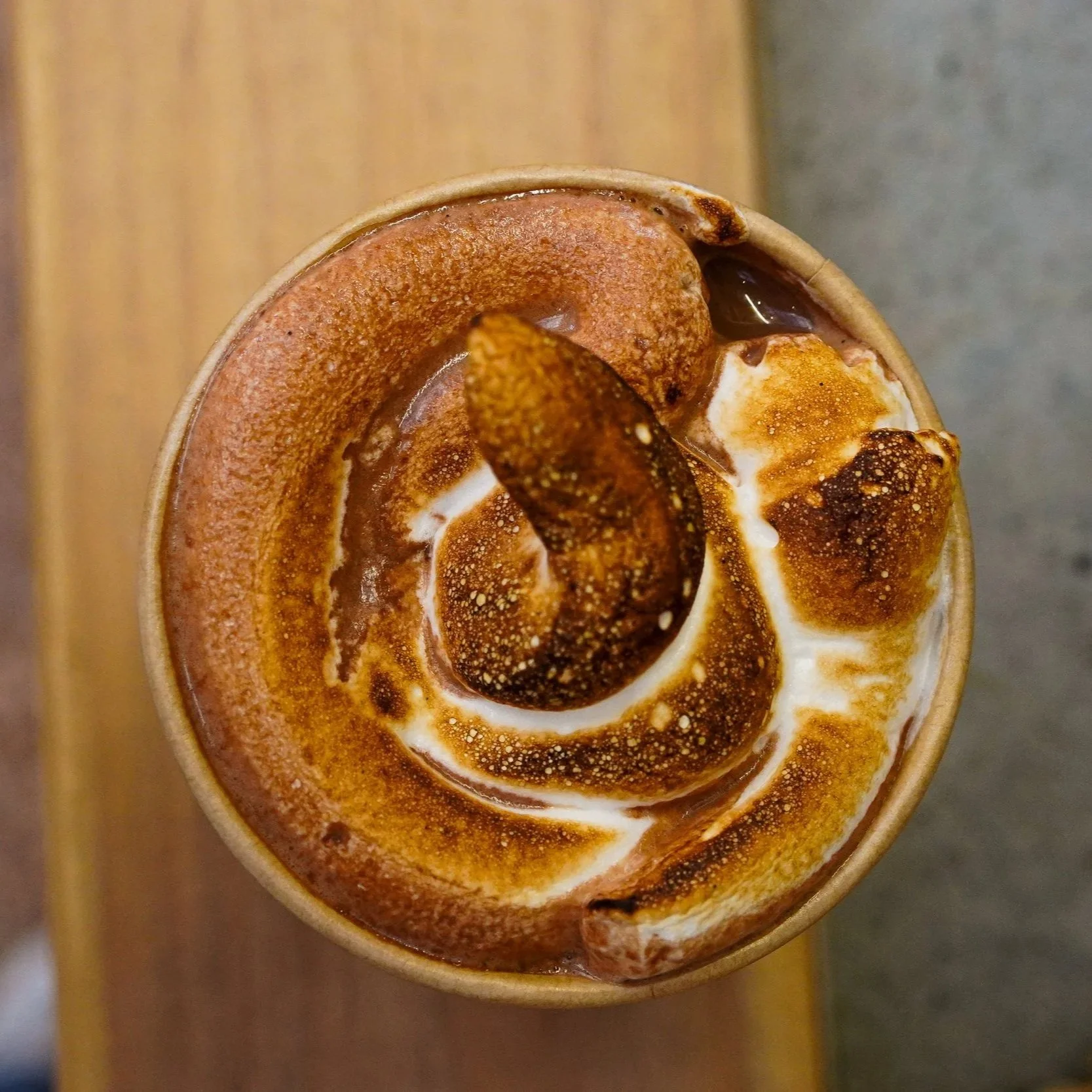 Close-up of a cinnamon roll with white icing and a chocolate-covered marshmallow on top, served in a paper cup on a wooden surface.