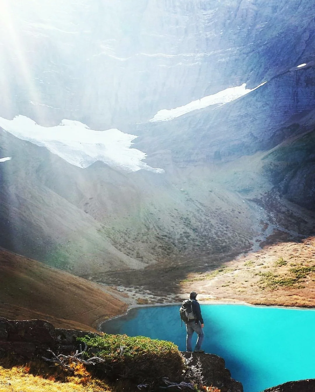 A hiker with a backpack standing near a bright blue mountain lake, surrounded by rocky and grassy terrain, with snow-capped mountains in the background.