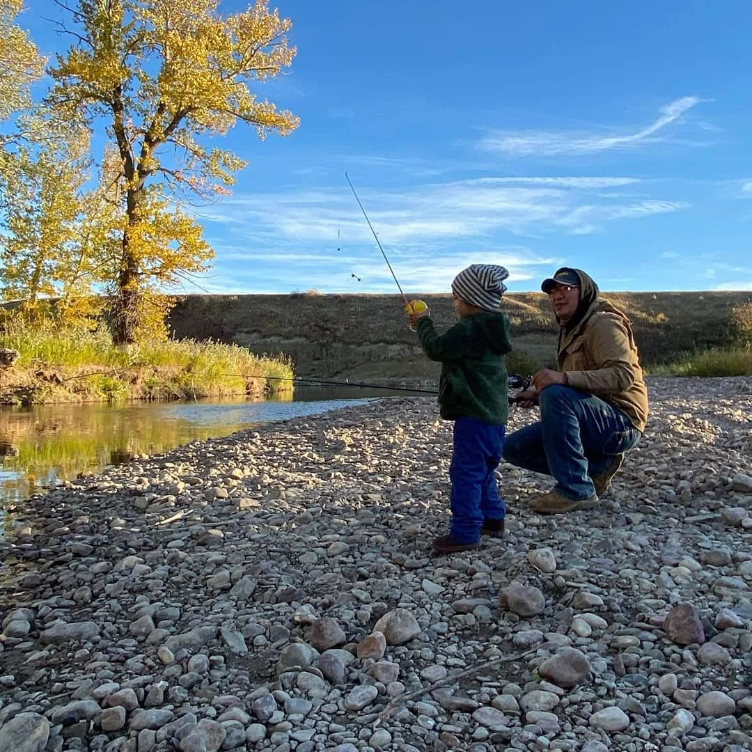 An adult and a child fishing together on a rocky riverbank with a tree and blue sky in the background.