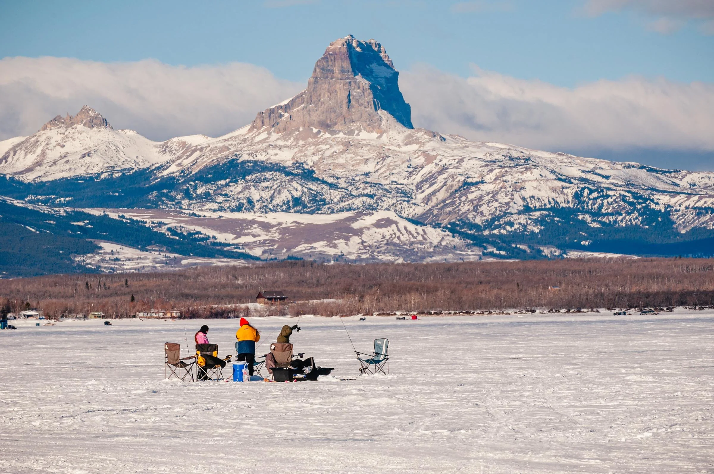 People sitting on chairs ice fishing on a frozen lake with snow-covered mountains and a prominent pinnacled mountain in the background.